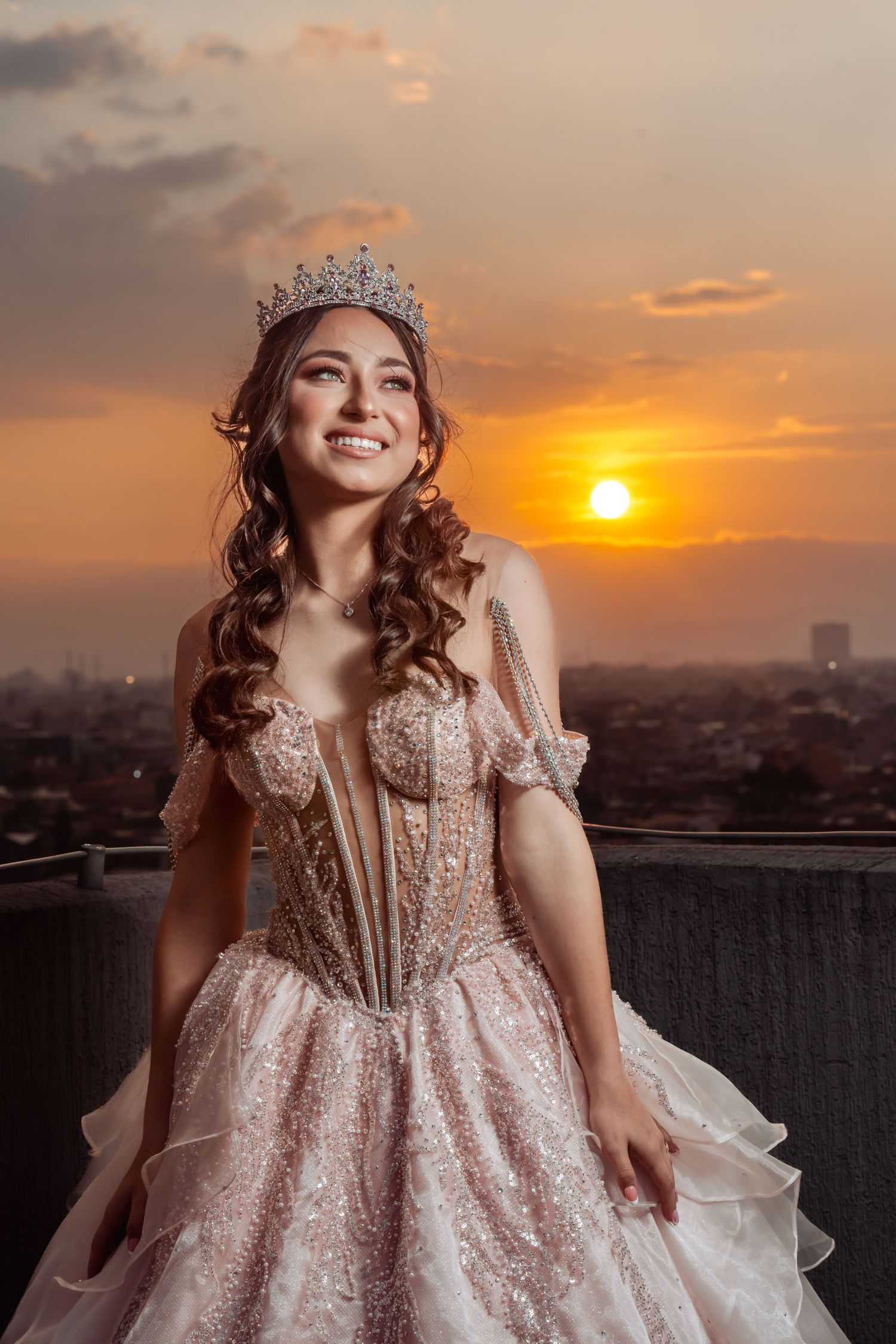 Retrato quinceañera sonriente con vestido rosa brillante (corsé) y tiara en terraza; sol poniéndose sobre ciudad y atardecer naranja.