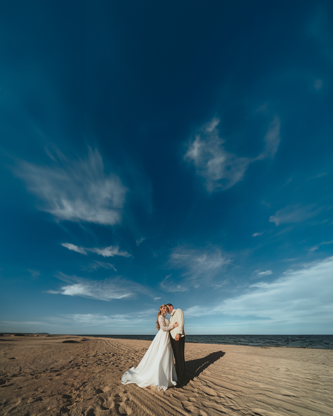 Novios se abrazan en playa de Miami bajo amplio cielo azul con nubes blancas. Fotografía boda destino.