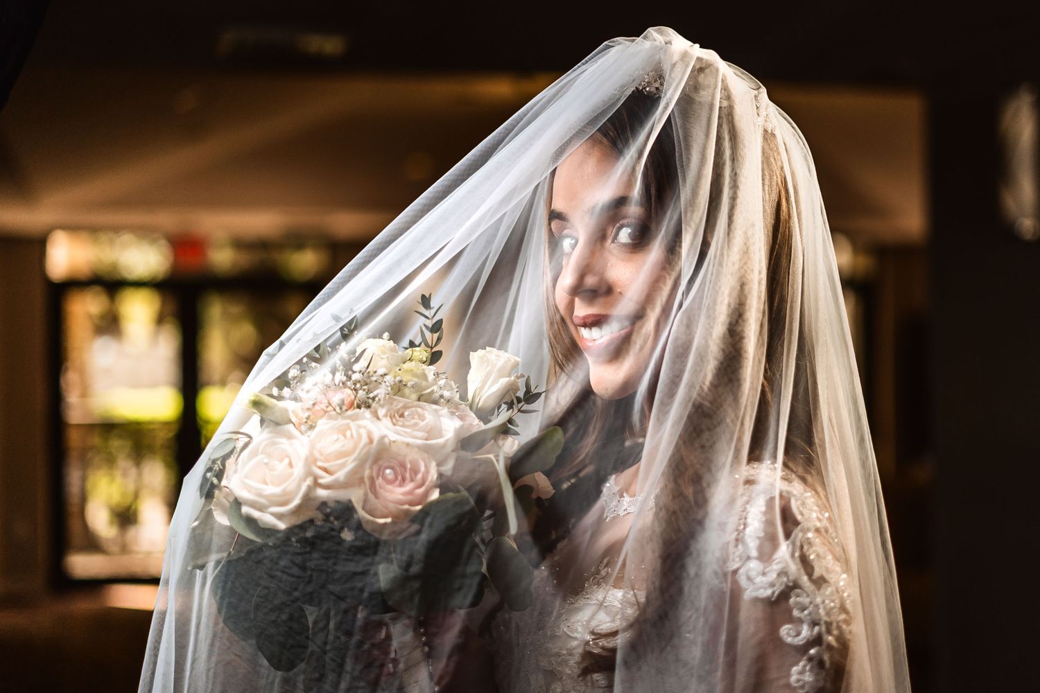 Retrato artístico novia con ramo mira a través de su velo etéreo; luz suave y misteriosa. Boda.
