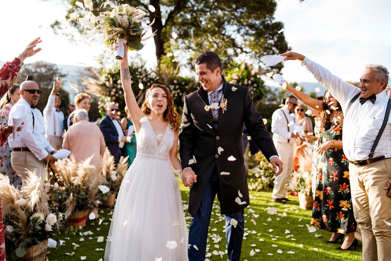 Radiante pareja recién casada caminando entre invitados que lanzan pétalos blancos. Novia con ramo elevado, vestido blanco novio traje oscur