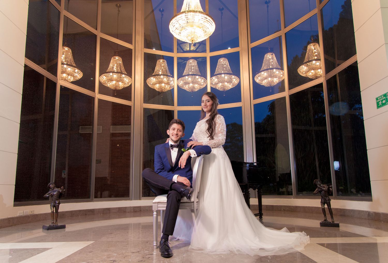 Novios posan en salón elegante; él sentado (traje azul), ella de pie (vestido blanco). Candelabros, piano. Boda.