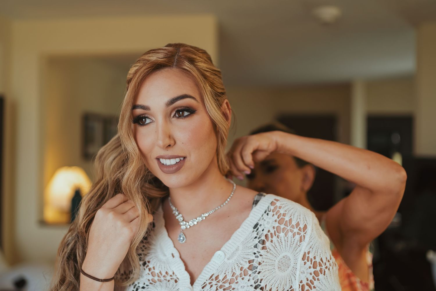 Novia con blusa de encaje recibe ayuda para colocar collar de diamantes; preparación boda, tocado.
