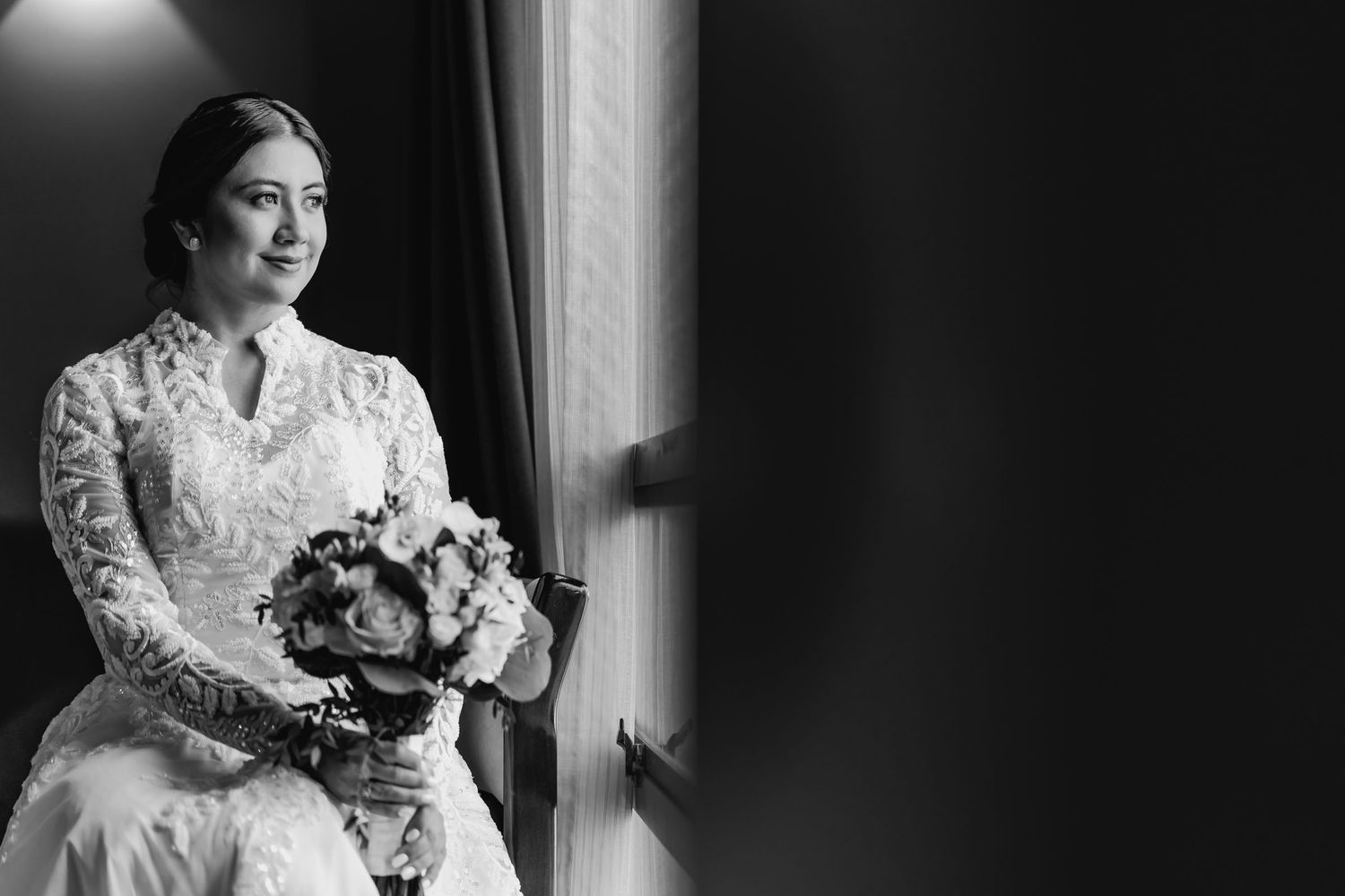 Foto blanco y negro: Novia con vestido de encaje y ramo mira por la ventana; retrato elegante durante preparación de boda.