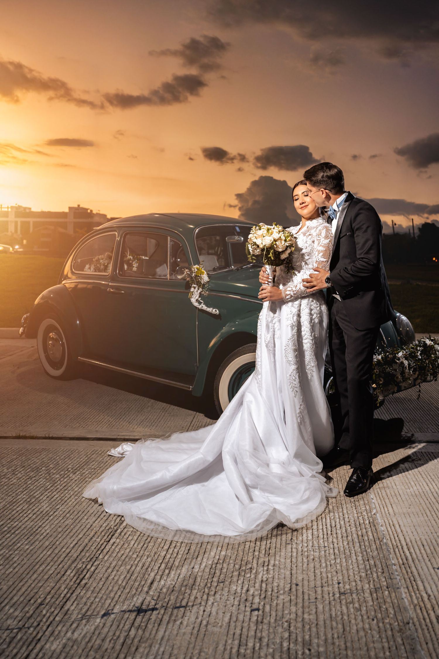Novios tomados de la mano miran con cariño junto a coche verde clásico al atardecer. Abrazo romántico en boda.