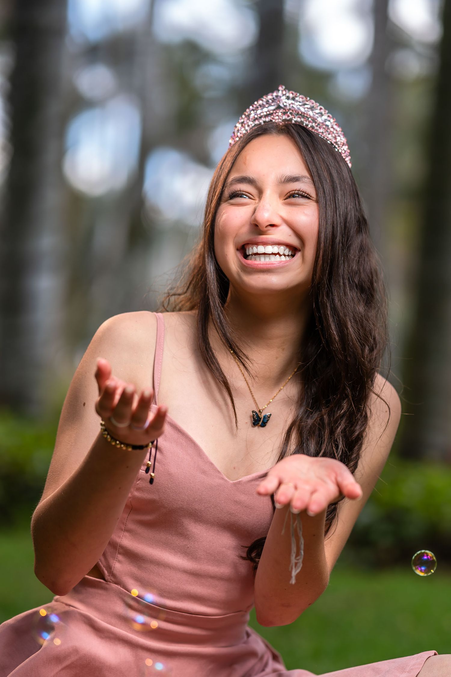Retrato: Joven ríe feliz con vestido rosa y tiara, extiende manos jugando con burbujas de jabón en bosque. Sesión fotos quinceañera.