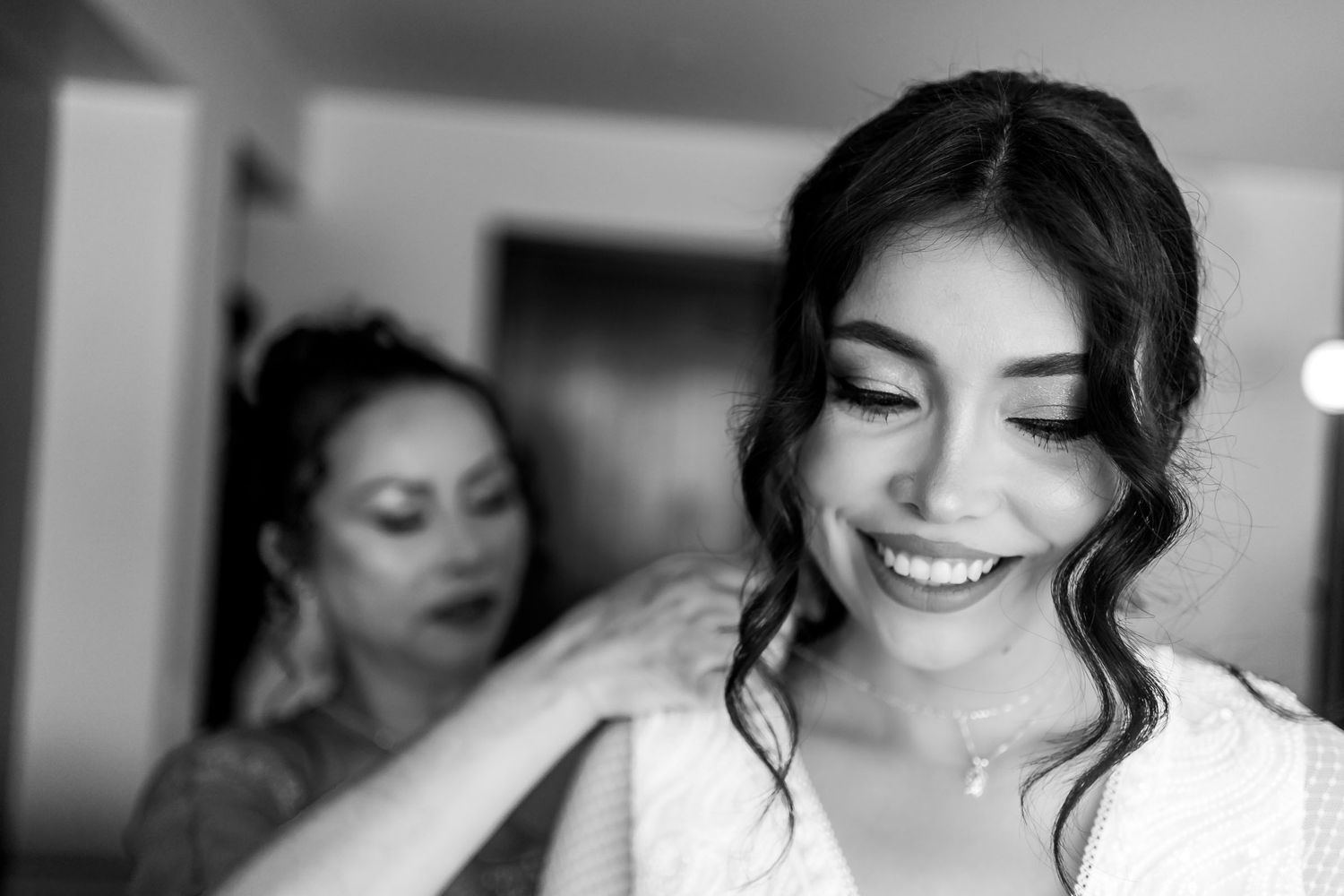 Primer plano blanco y negro de novia sonriendo con ojos cerrados mientras mujer ajusta su vestido. Momento de preparación boda.