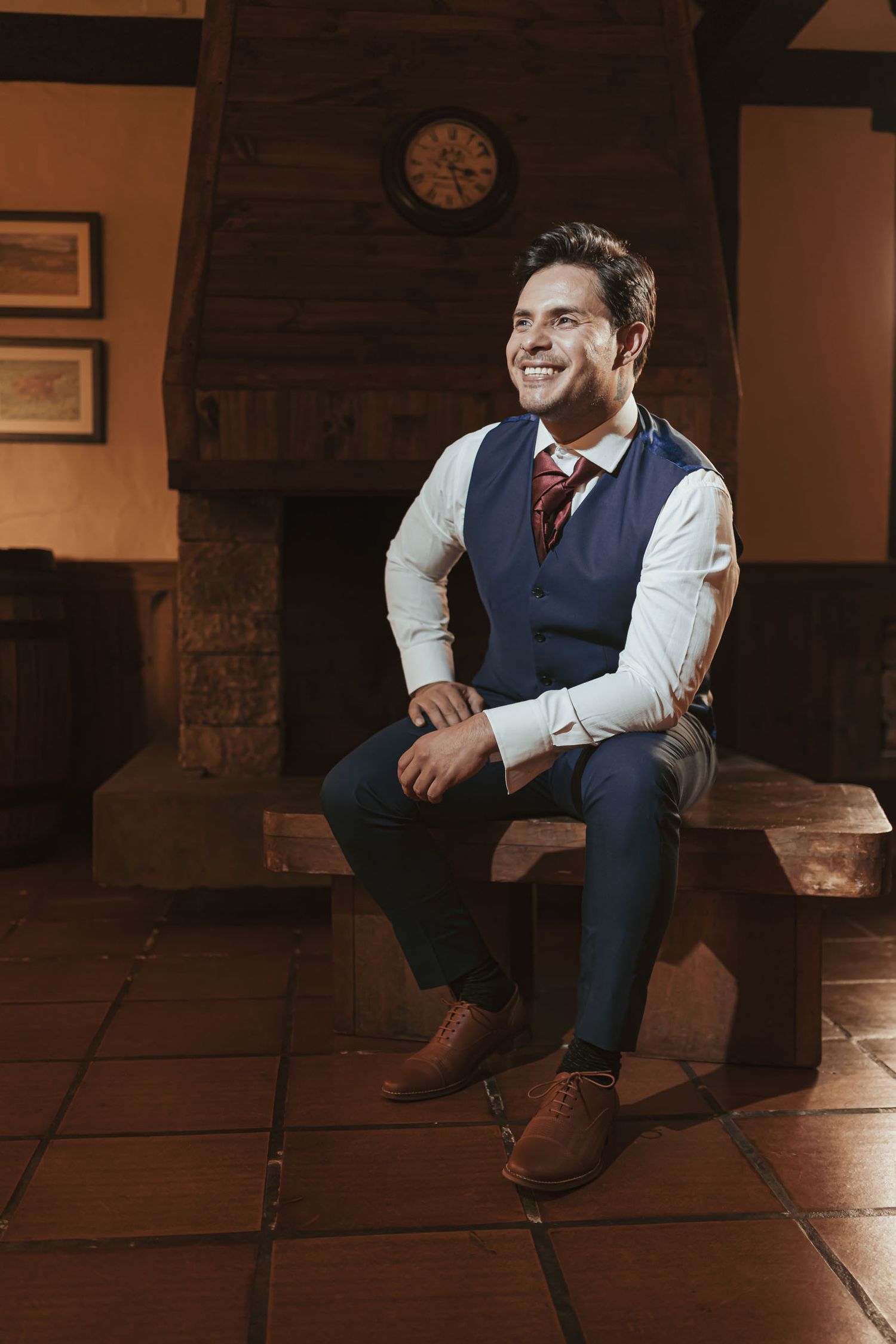 Hombre elegante con chaleco azul y corbata, sentado en banco madera frente a chimenea; interior rústico. Alistamiento novio boda