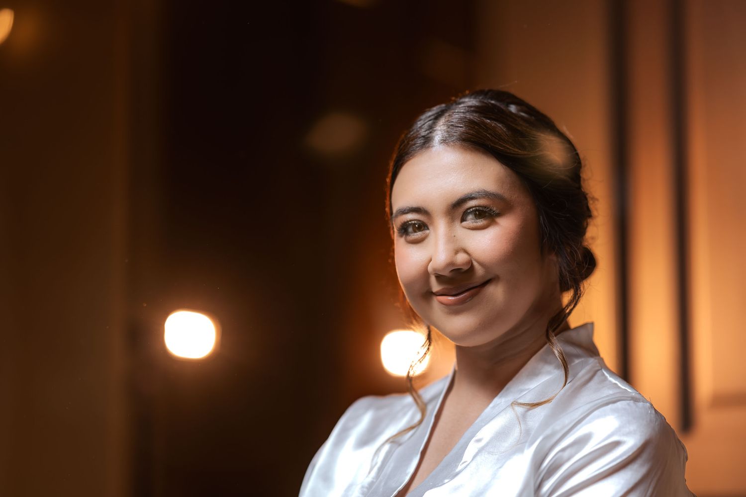 Retrato de novia sonriente con bata de satín blanca; luces cálidas desenfocadas al fondo. Preparación boda.
