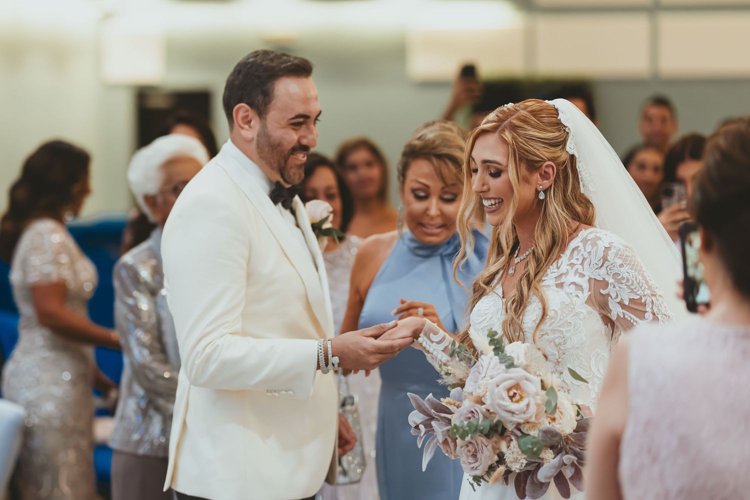 Novio (traje blanco) coloca anillo en dedo de novia sonriente; madre mira. Ceremonia boda.