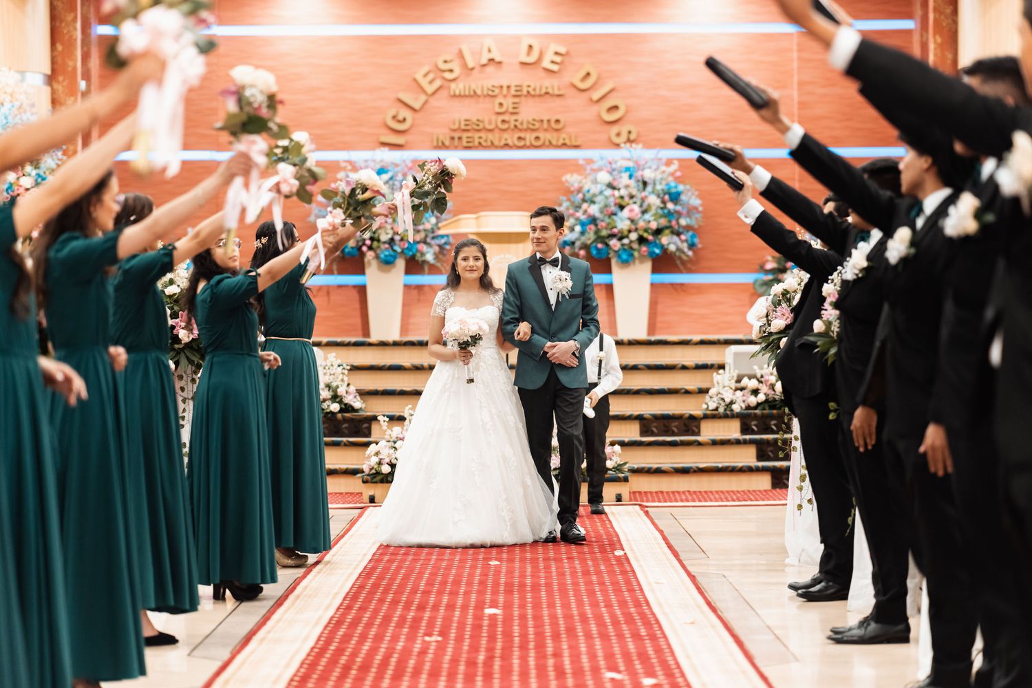 Pareja de recién casados desfilando bajo arco de biblias y flores en emotiva salida de iglesia.