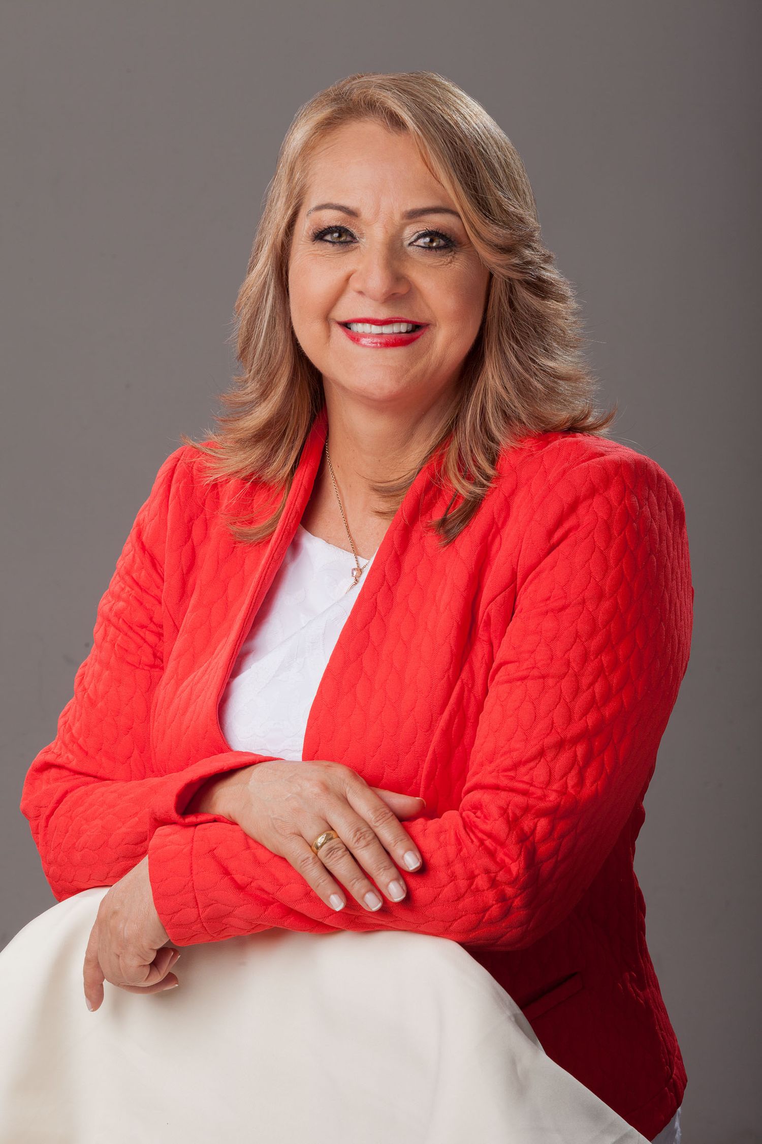 Retrato mujer sonriente con blazer rojo texturizado y top blanco, apoyada; fondo gris estudio. Headshot. Aydeé Lizarazo