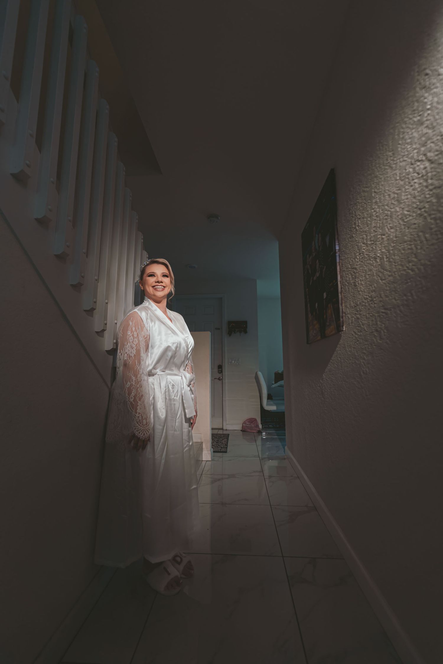 Novia con bata blanca posa sonriente en pasillo tenue cerca de escaleras; preparación boda.