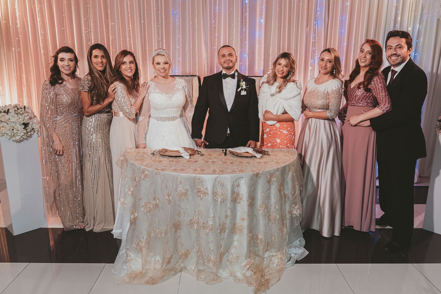 Foto grupal boda: Novios posan con familiares o cortejo tras mesa decorada; fondo cortina luces.