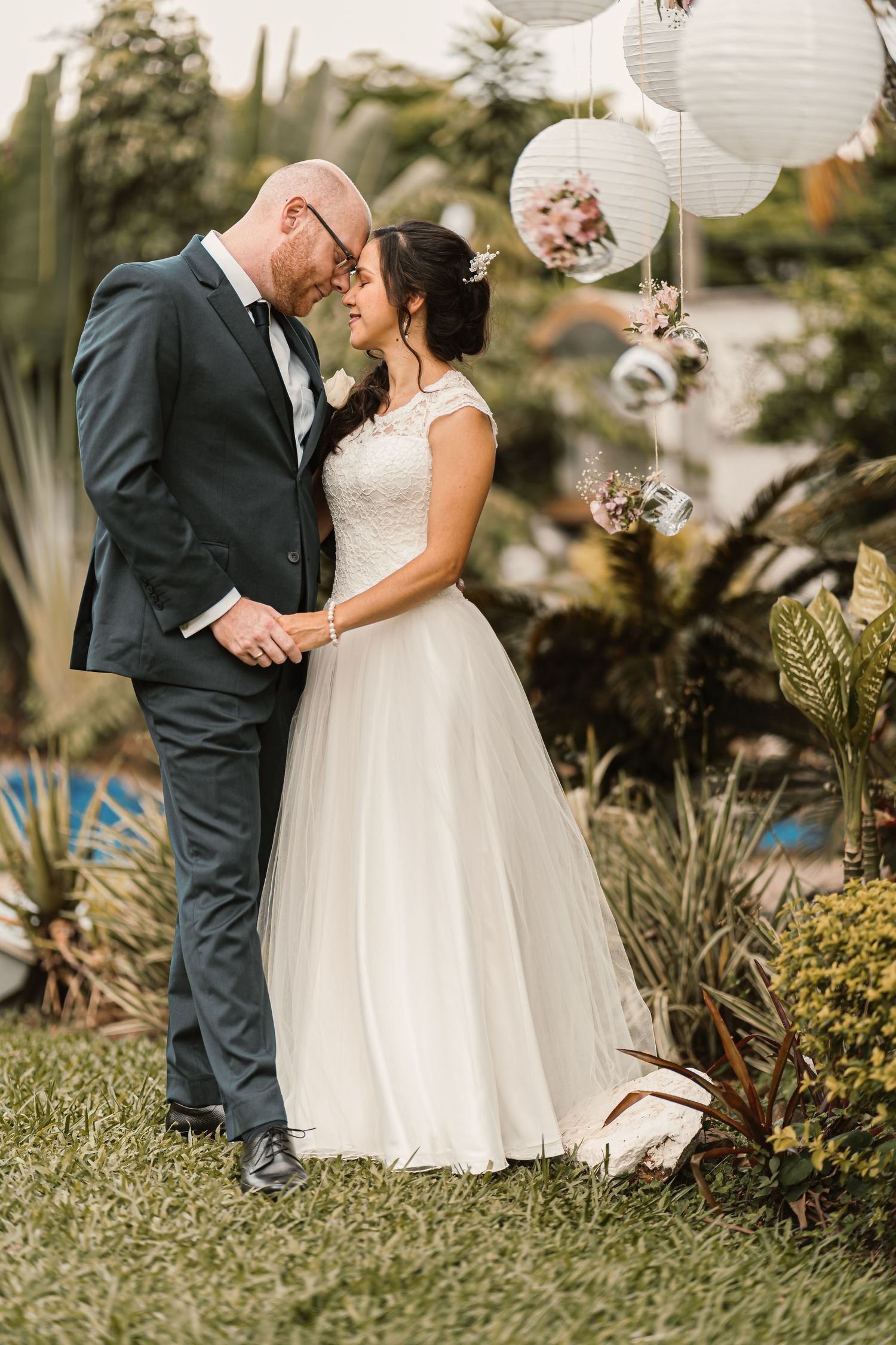 Novios se miran íntimamente (frentes juntas) en jardín; ella vestido blanco sencillo. Boda romántica.