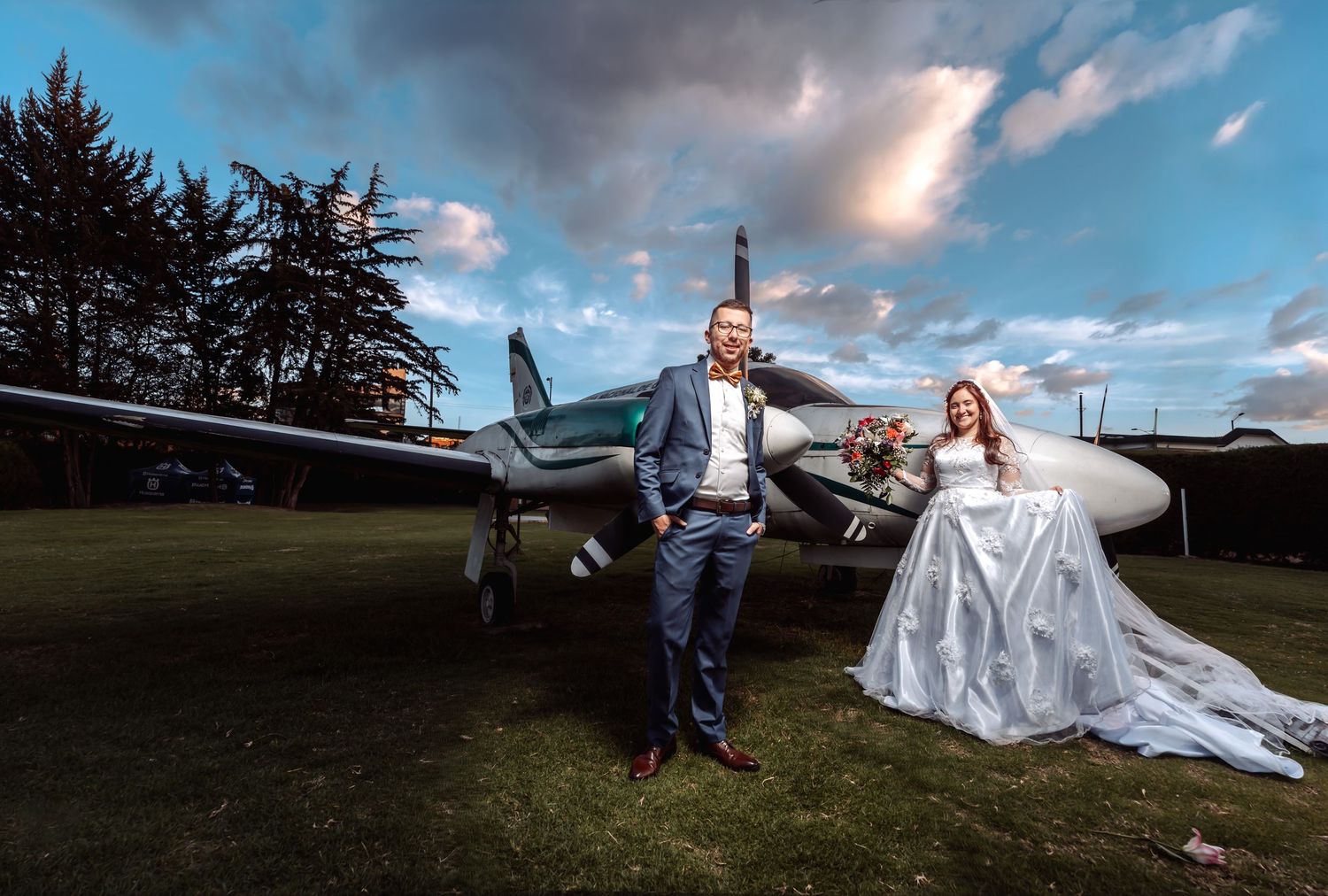 Novios posan elegantes junto a avioneta plateada en campo; novia con velo largo. Boda temática.