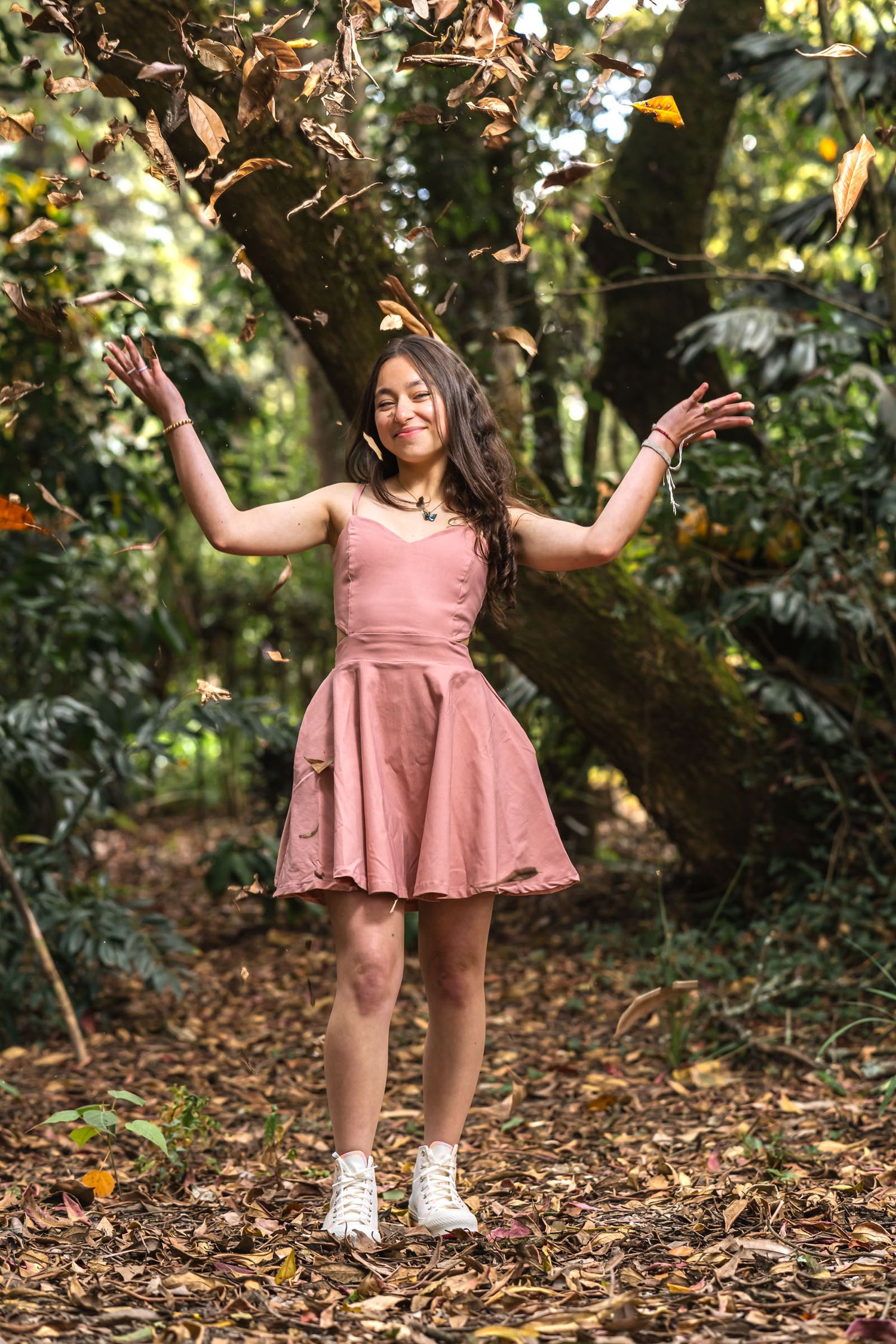 Joven con vestido corto rosa y zapatillas blancas lanza hojas secas al aire en bosque; sesión prequince.