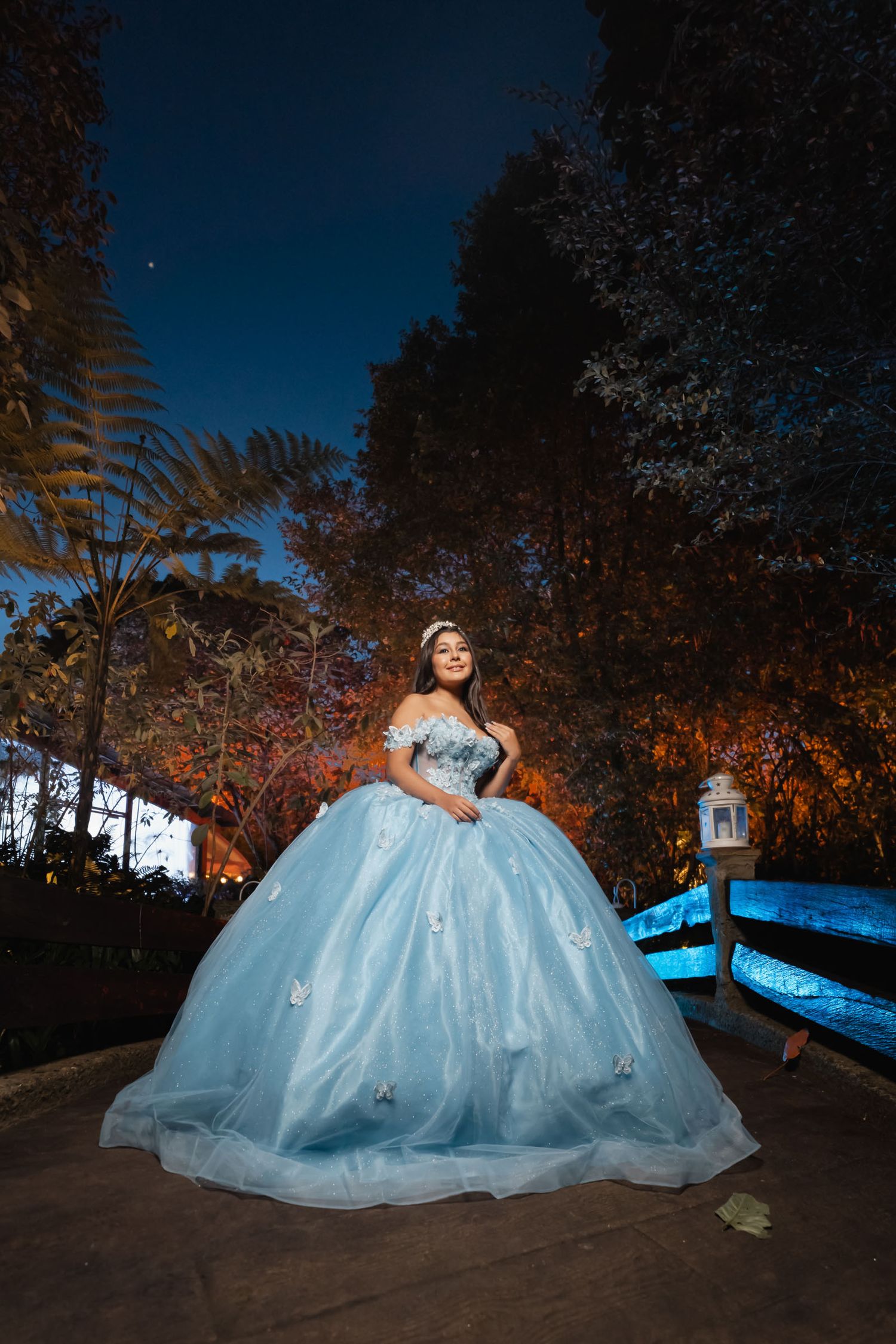 Joven quinceañera con vestido azul princesa y tiara posa de noche en puente; cielo estrellado. Mágico.