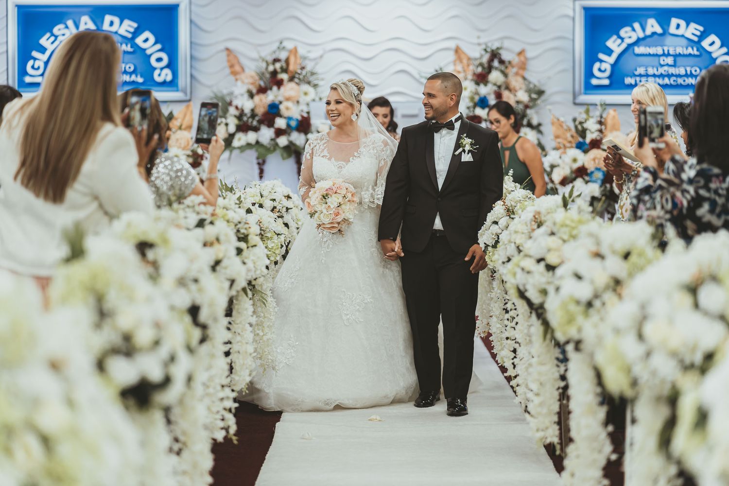 Salida nupcial: Novios caminan por pasillo iglesia (Iglesia de Dios); invitados graban con móviles. Boda IDMJI.