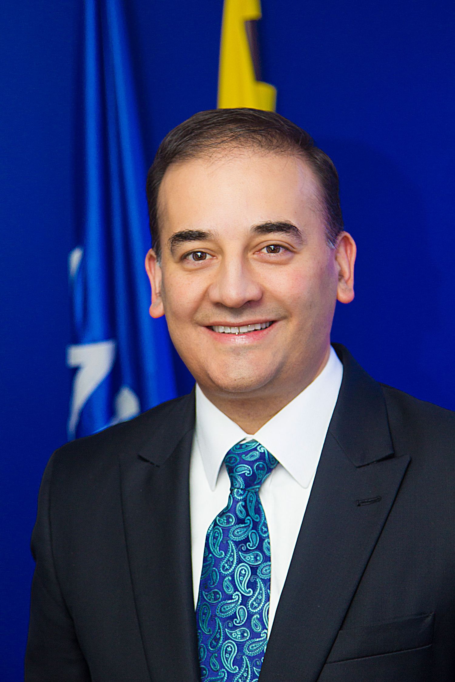 Retrato hombre sonriente con traje oscuro y corbata azul estampada; fondo banderas azules. Headshot oficial. Carlos Alberto Baena