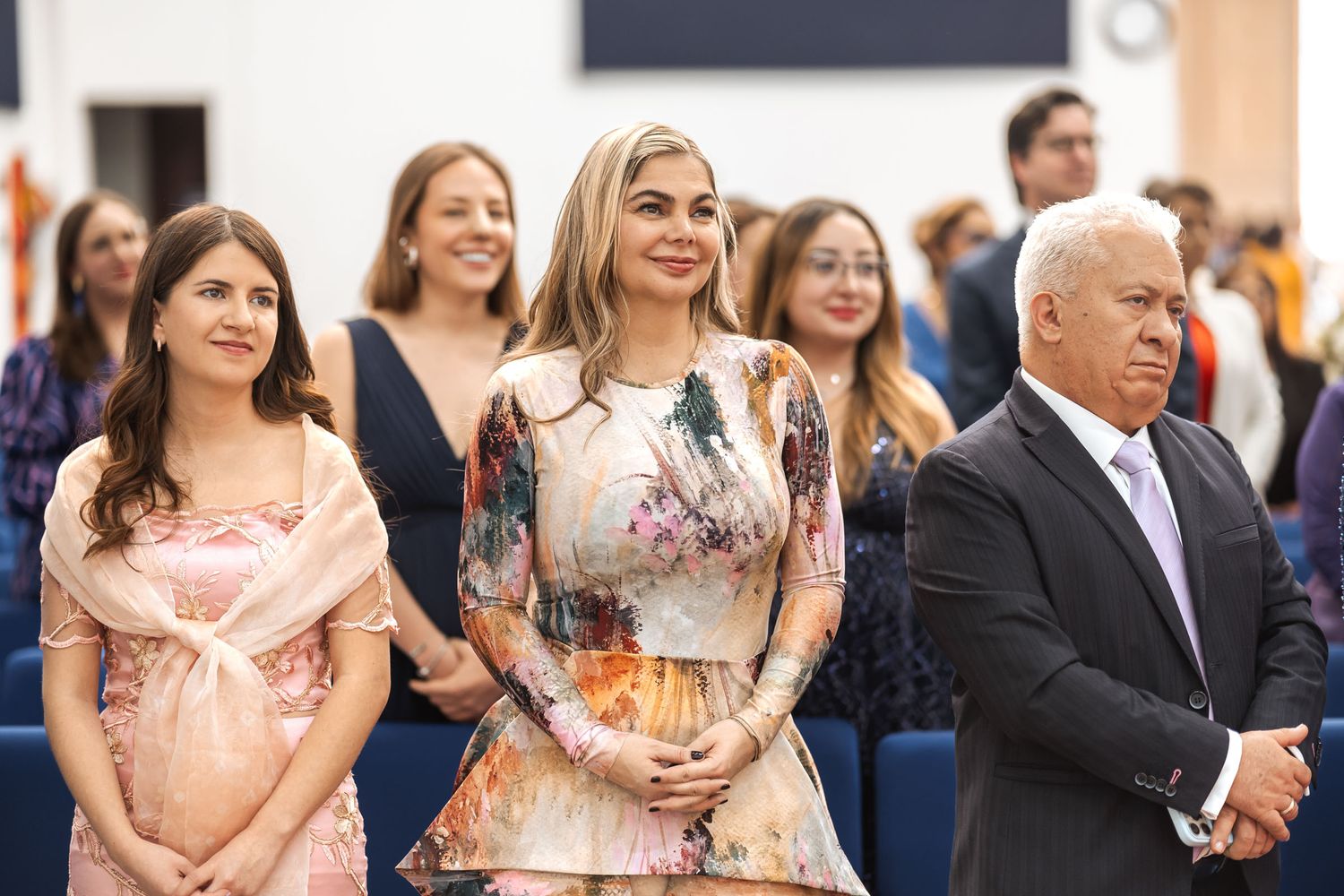Invitados atentos durante ceremonia de boda sentados en iglesia; mujer con vestido floral colorido en primer plano.