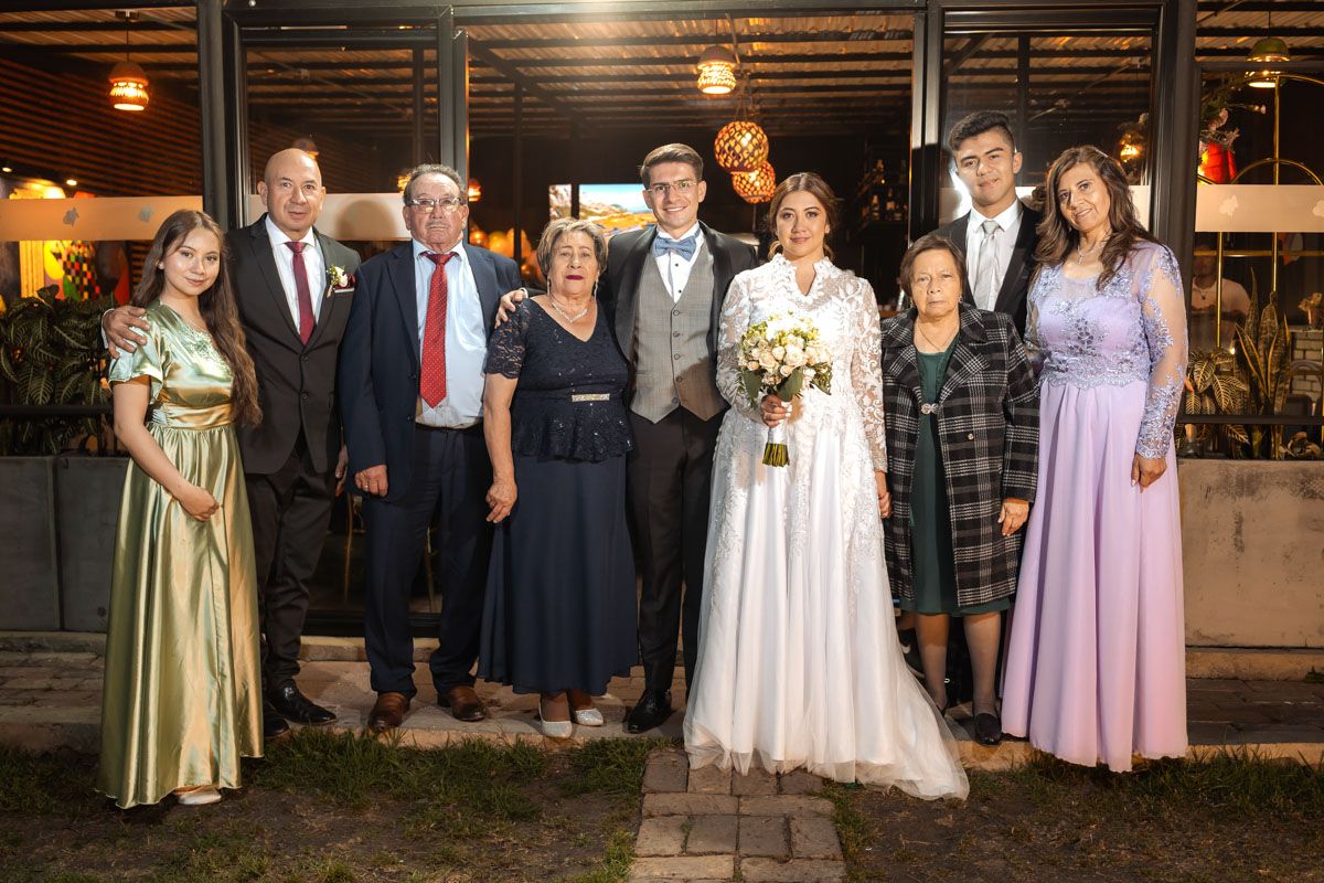 Novios posan felices con su familia, incluyendo niña, en foto de grupo exterior de noche. Boda.