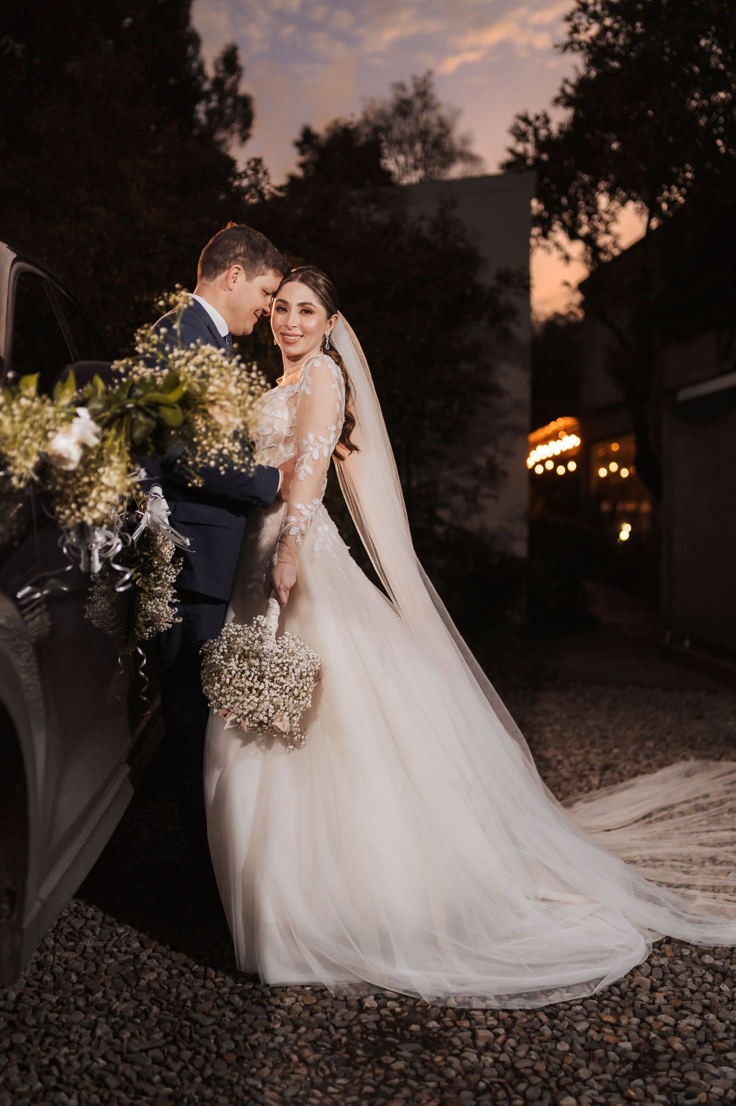 Novios posan junto a coche decorado con flores al atardecer; ella sostiene ramo, él traje azul. Velo largo. Boda.