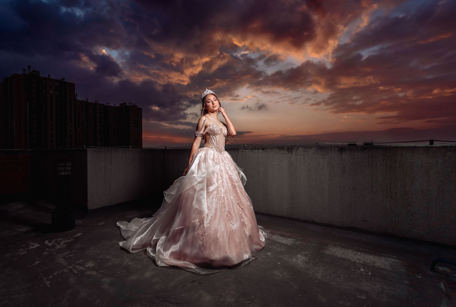Quinceañera en vestido y tiara posa en terraza con espectacular cielo al atardecer. Foto de celebración.