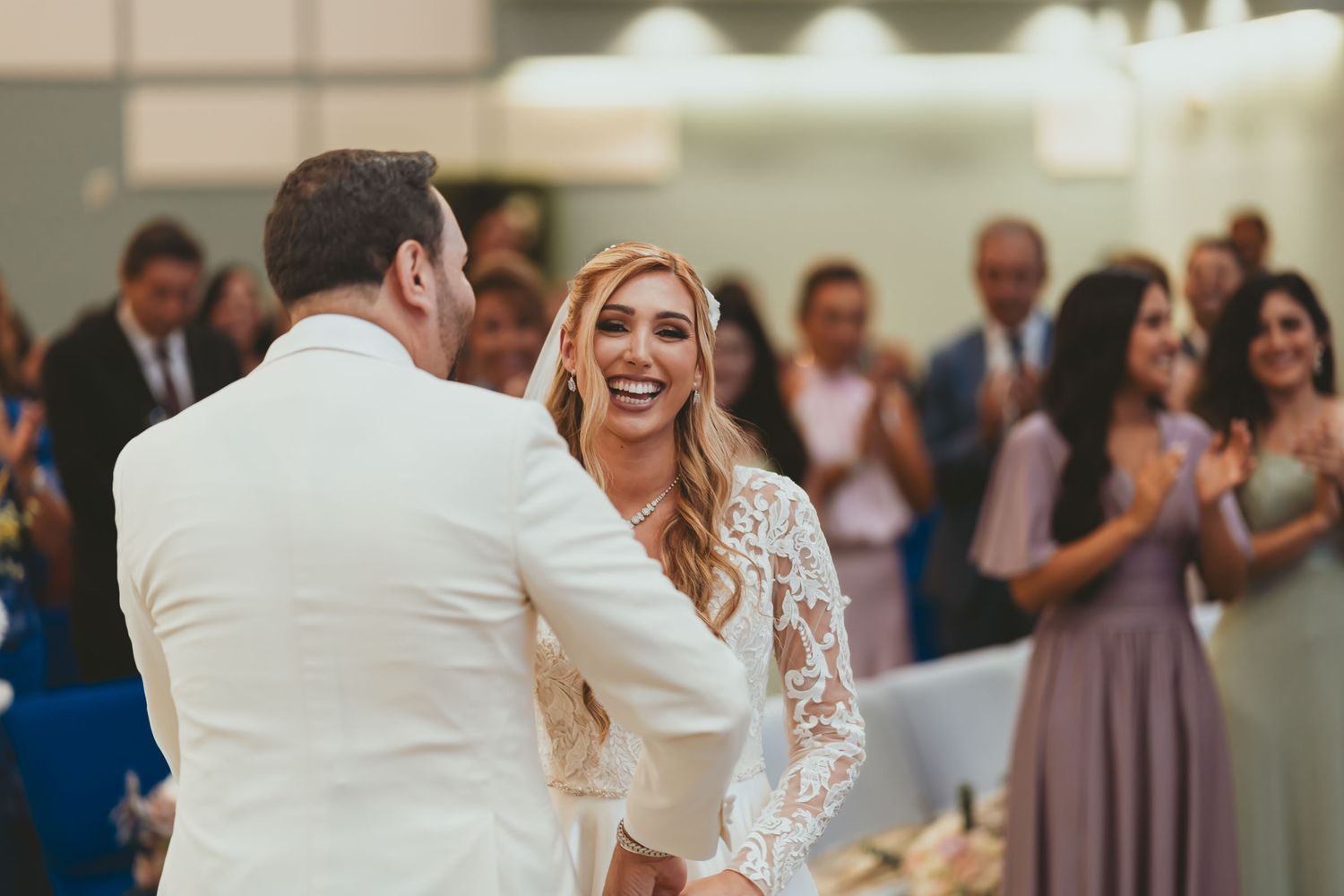 Novia ríe feliz abrazando al novio (chaqueta blanca) en recepción; invitados aplauden al fondo. Boda.