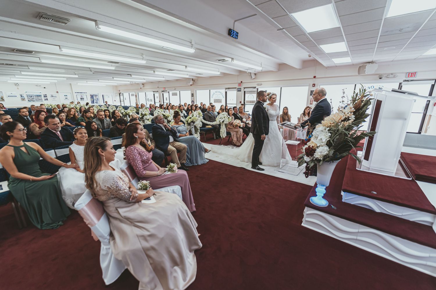 Vista amplia de ceremonia boda en salón/iglesia; novios y oficiante al frente, invitados sentados observan.