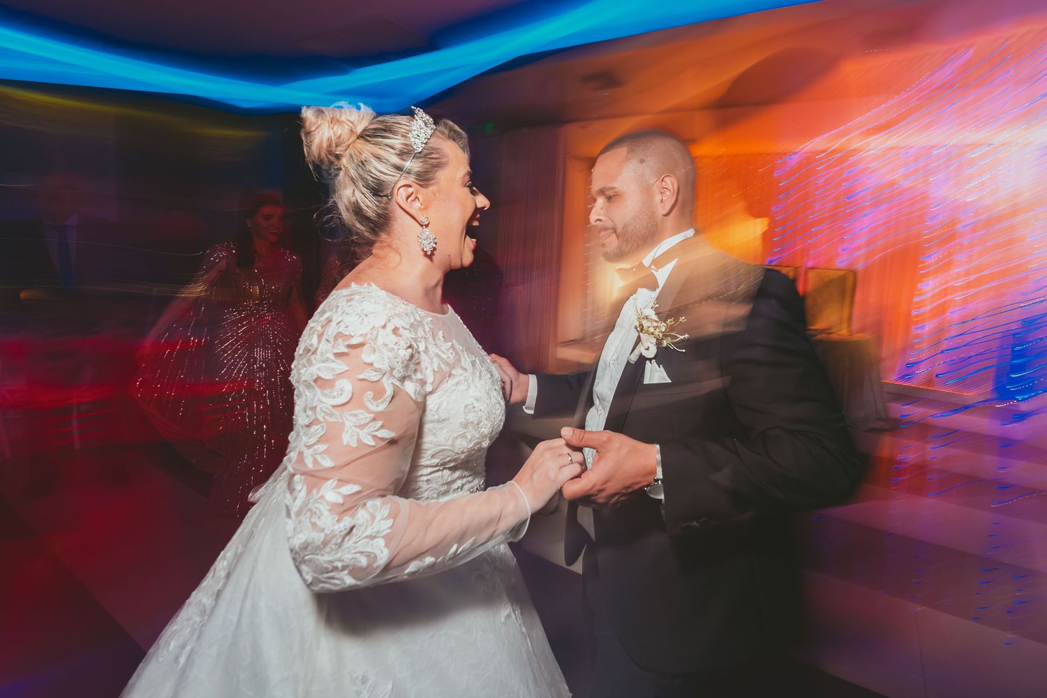 Novia y novio ríen mientras bailan juntos en recepción; foto con movimiento y luces de fiesta coloridas.