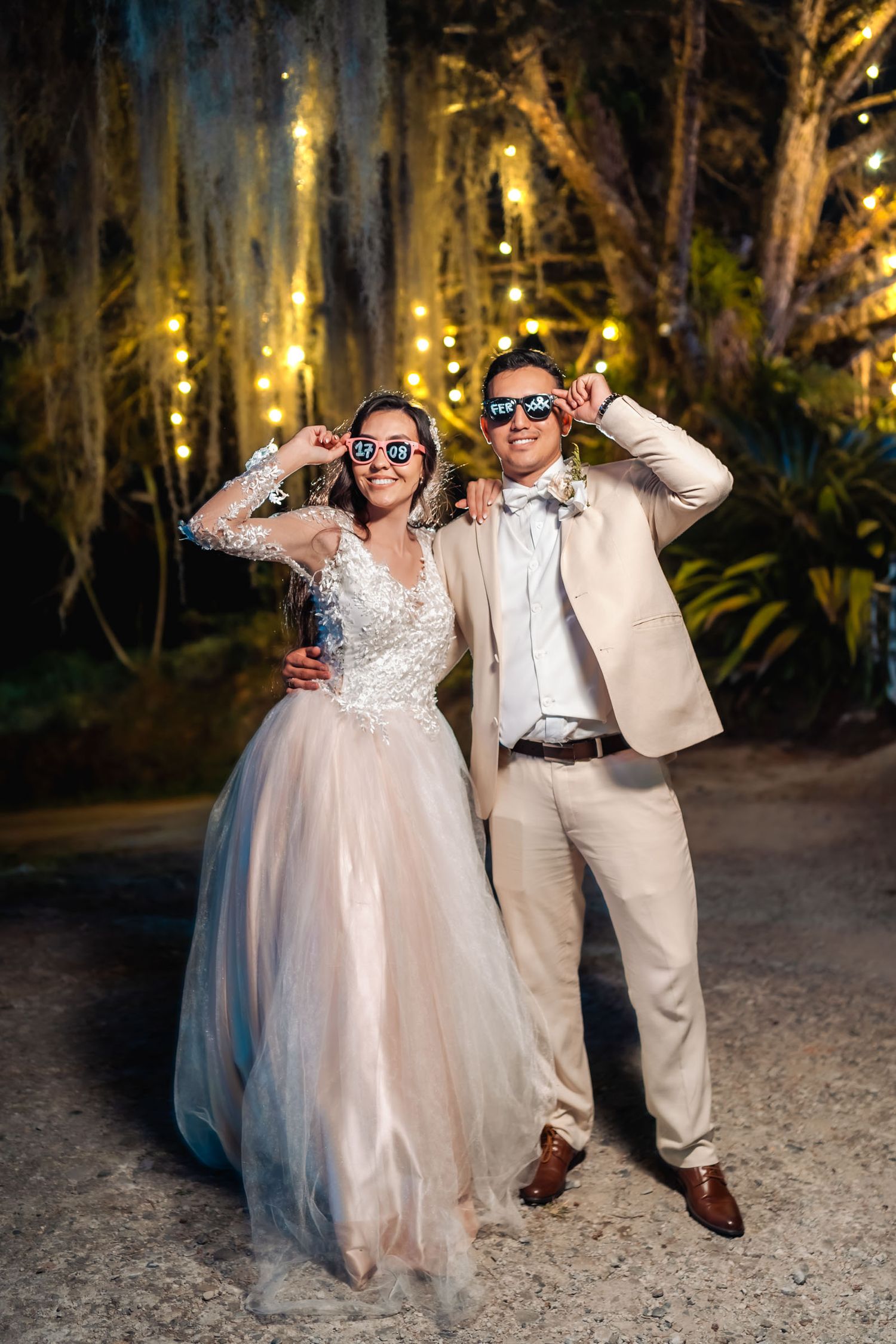 Novios posan divertidos con gafas de fiesta en recepción nocturna; luces cálidas fondo.