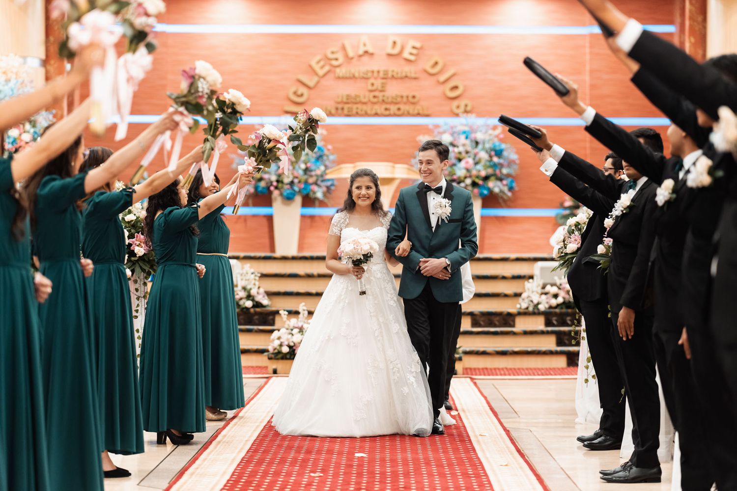 Boda religiosa: recién casados caminando por alfombra roja bajo arco de flores y saludos de invitados. Novia con vestido blanco, novio traje