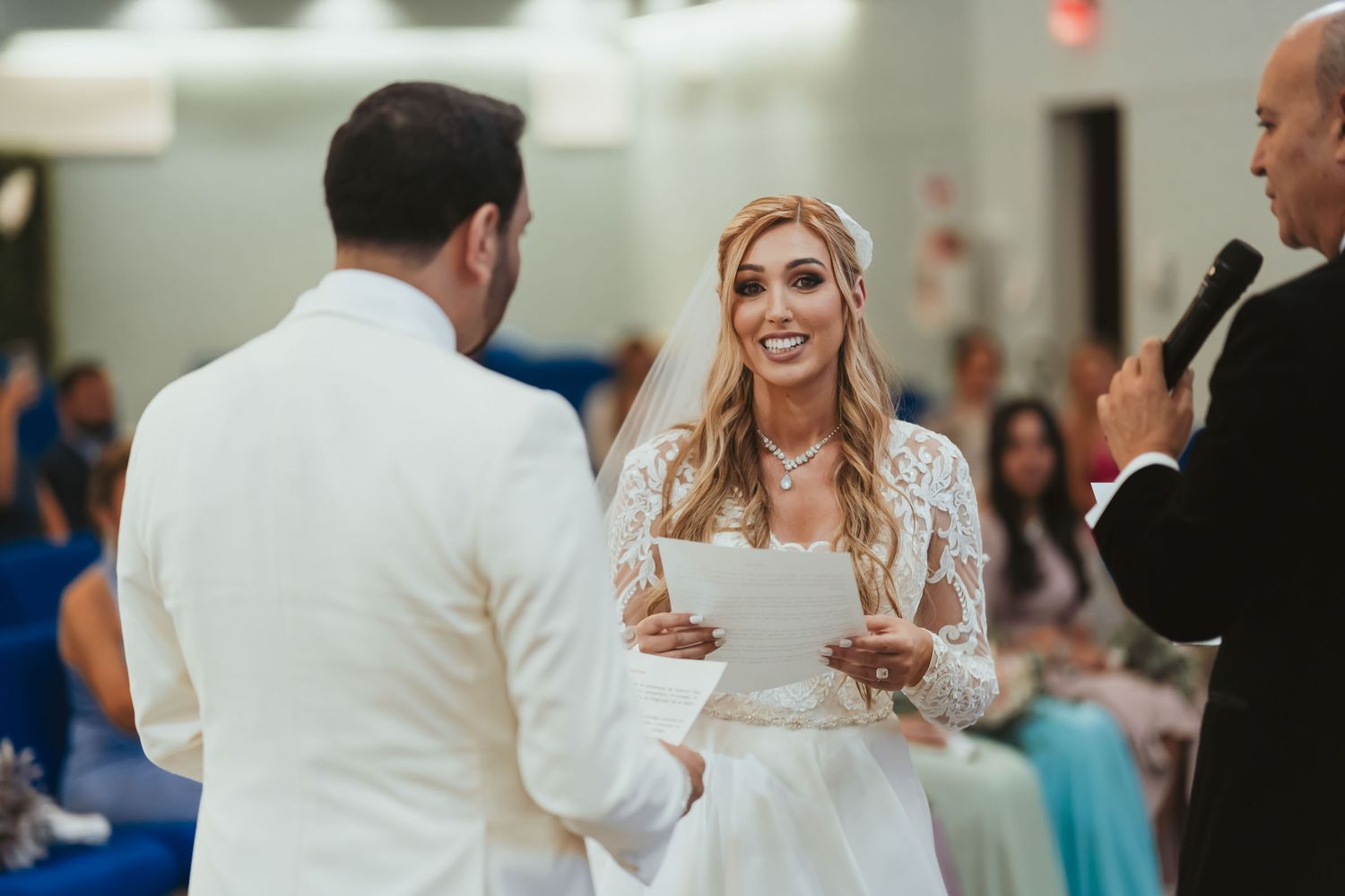 Novia sonriente lee sus votos al novio (traje blanco) durante ceremonia boda; oficiante observa.