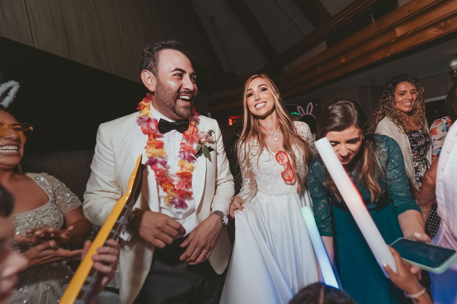 Novios con collares de flores bailan felices en fiesta 'hora loca' boda; invitados con glow sticks.