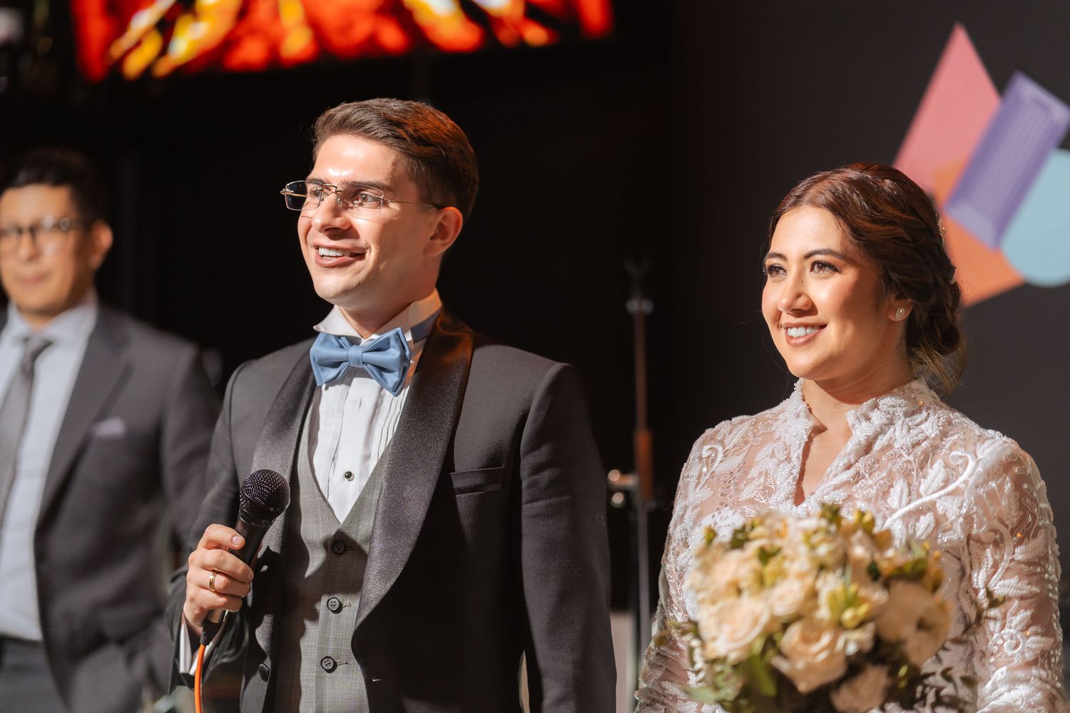 Novios sonríen en escenario de recepción de boda. Él sostiene micrófono, ella ramo. Foto de celebración.