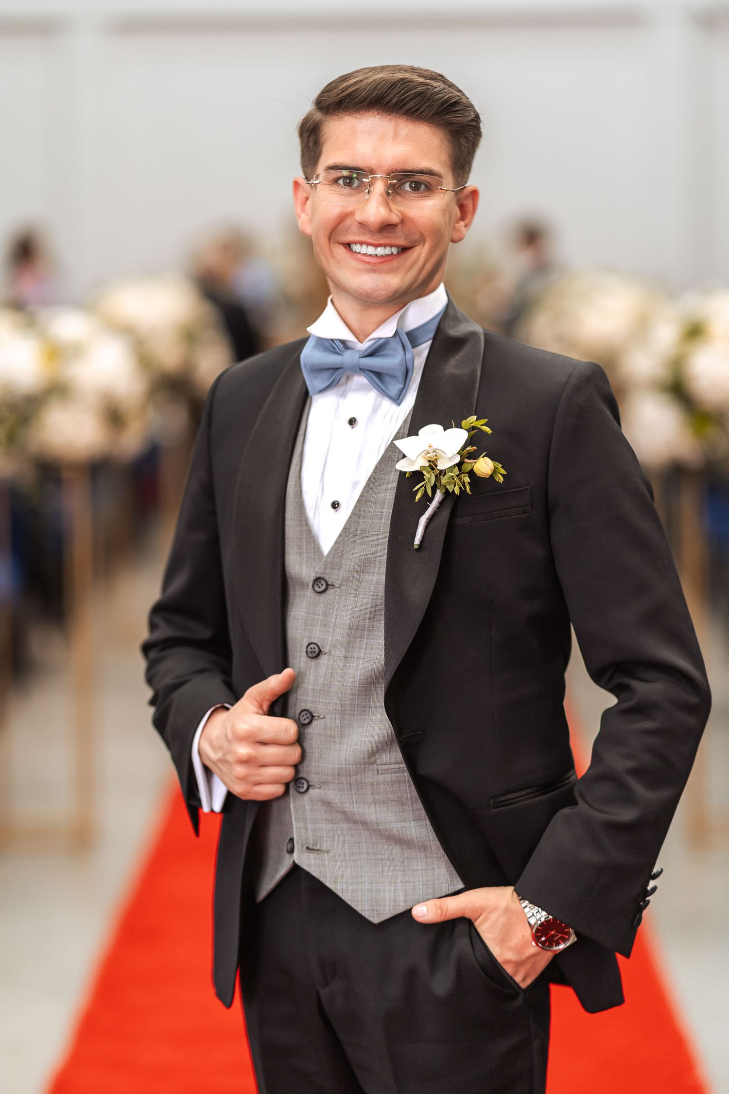 Novio sonriente con esmoquin, chaleco gris y pajarita azul posa en pasillo de iglesia sobre alfombra roja durante su boda.