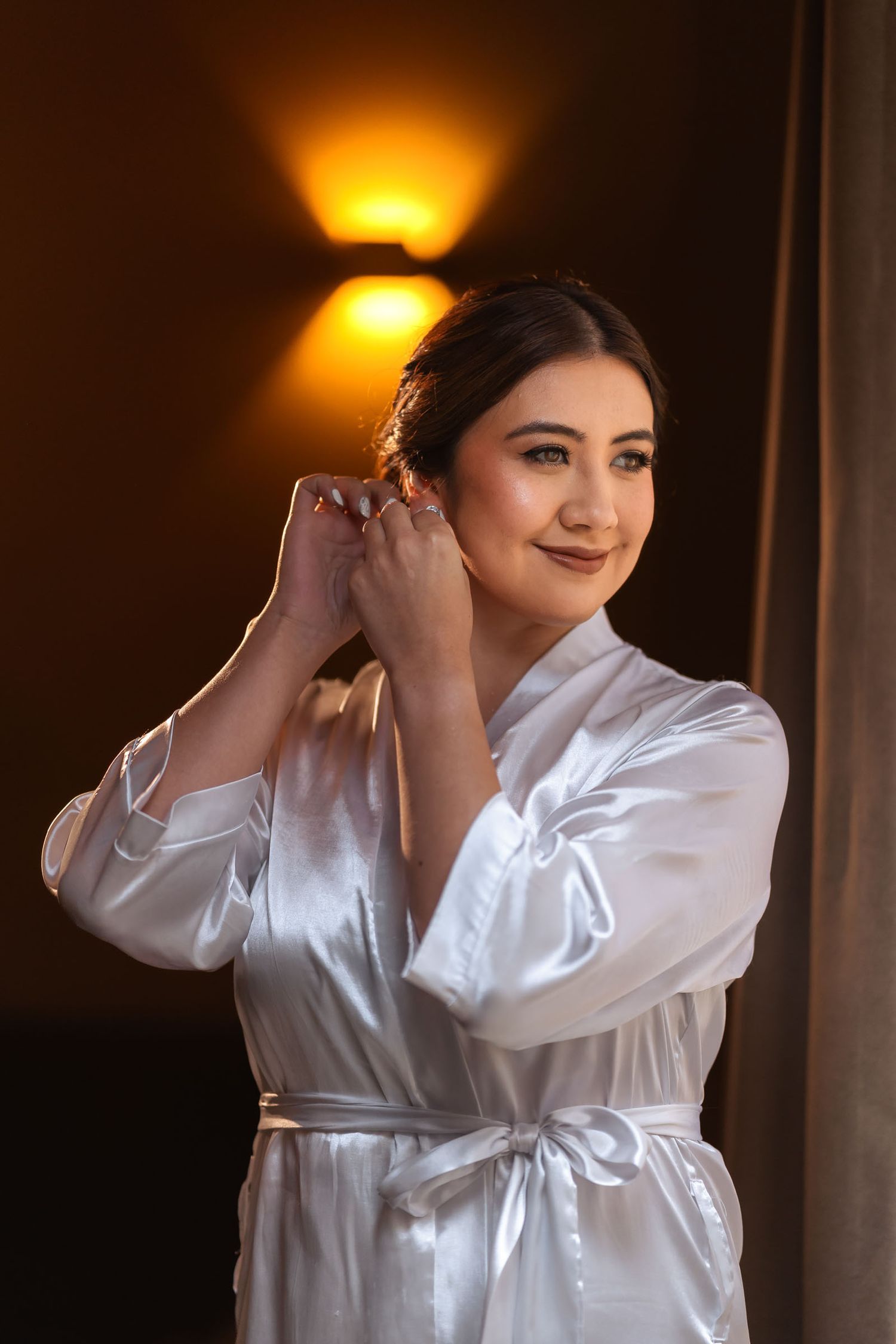 Novia con bata blanca de satín ajusta su arete sonriendo; preparación de boda, luz cálida de pared.