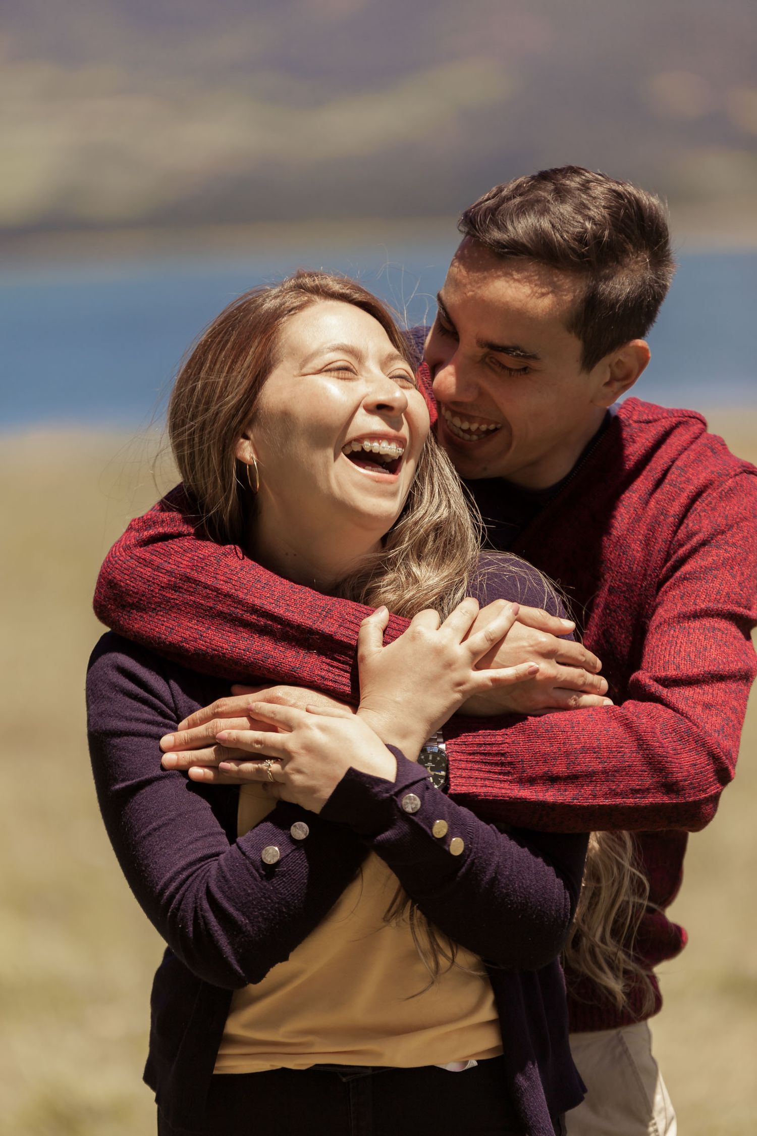 Pareja ríe felizmente abrazada al aire libre; hombre abraza por detrás a mujer, lago y montañas fondo.