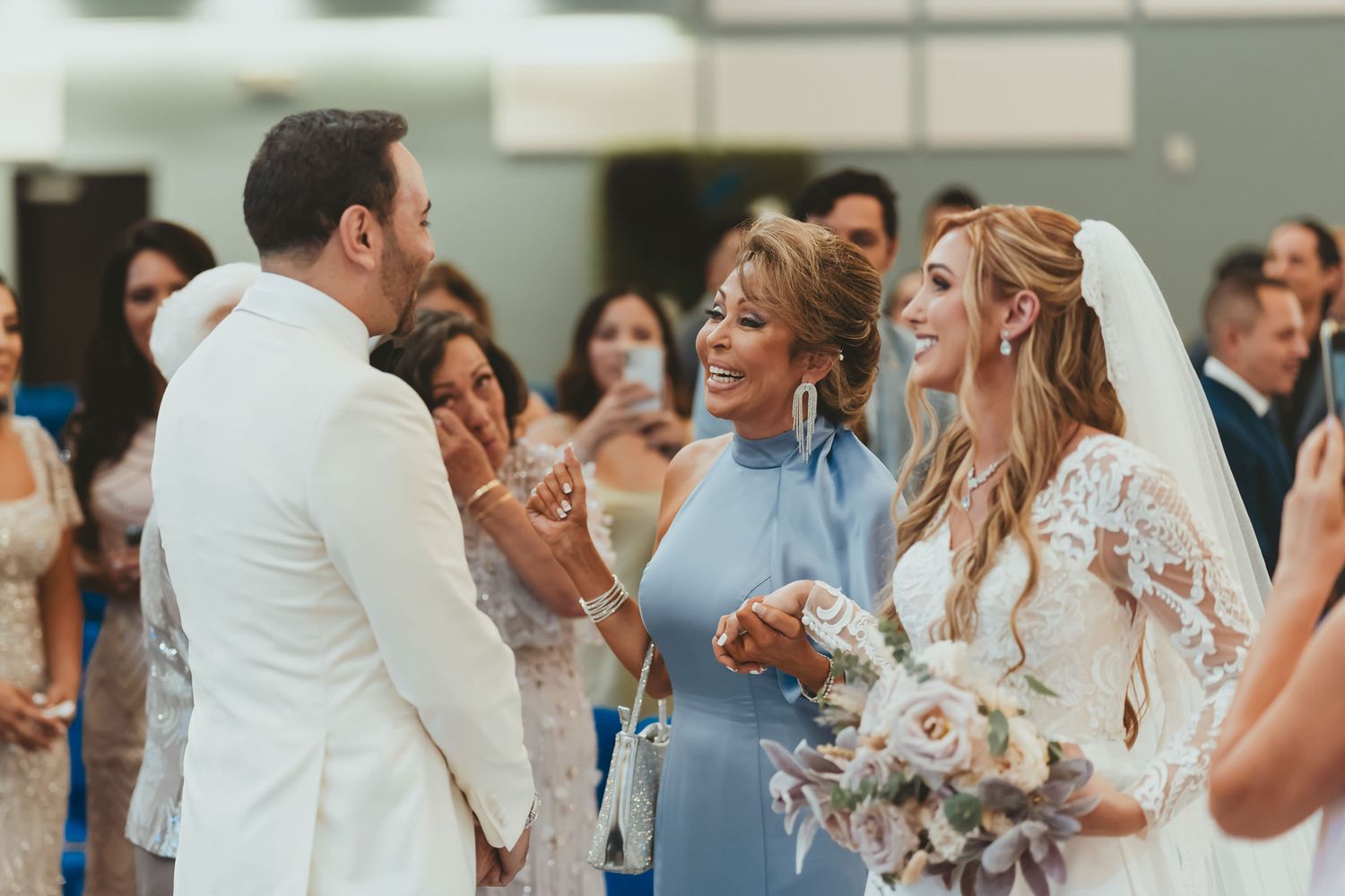 Novia y madre sonrientes miran al novio (traje blanco) en ceremonia boda; invitados fondo.