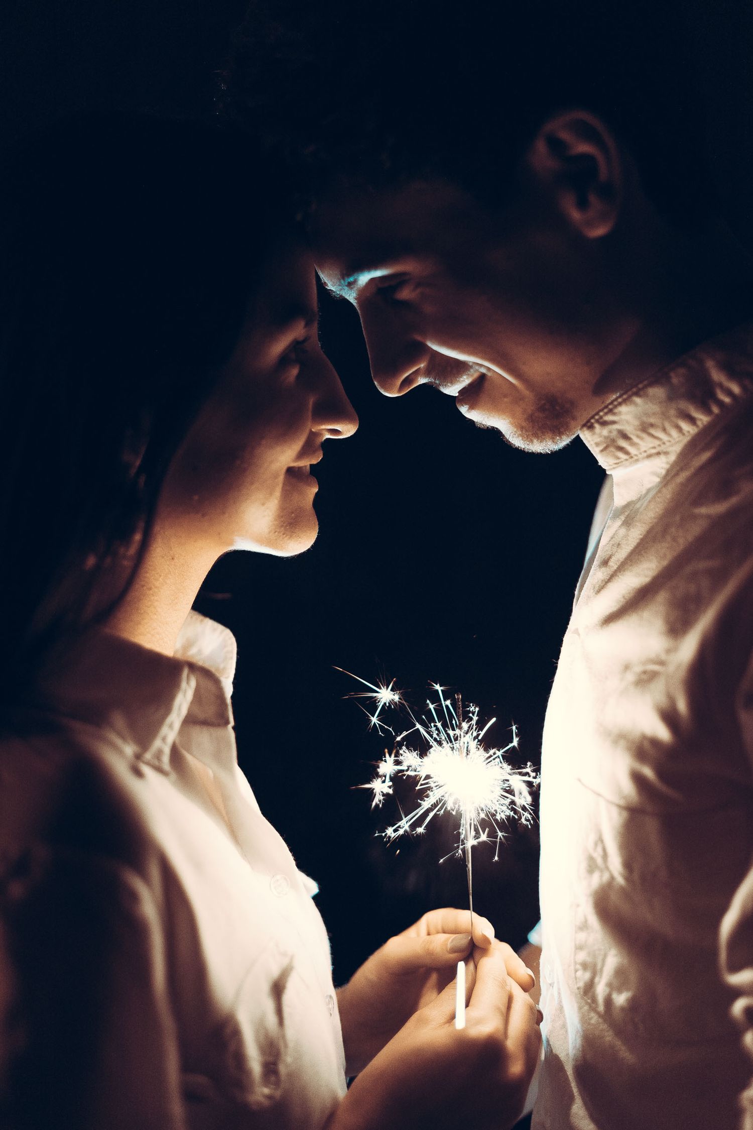 Pareja sonriente se mira de cerca sosteniendo bengala encendida en la oscuridad; momento íntimo.