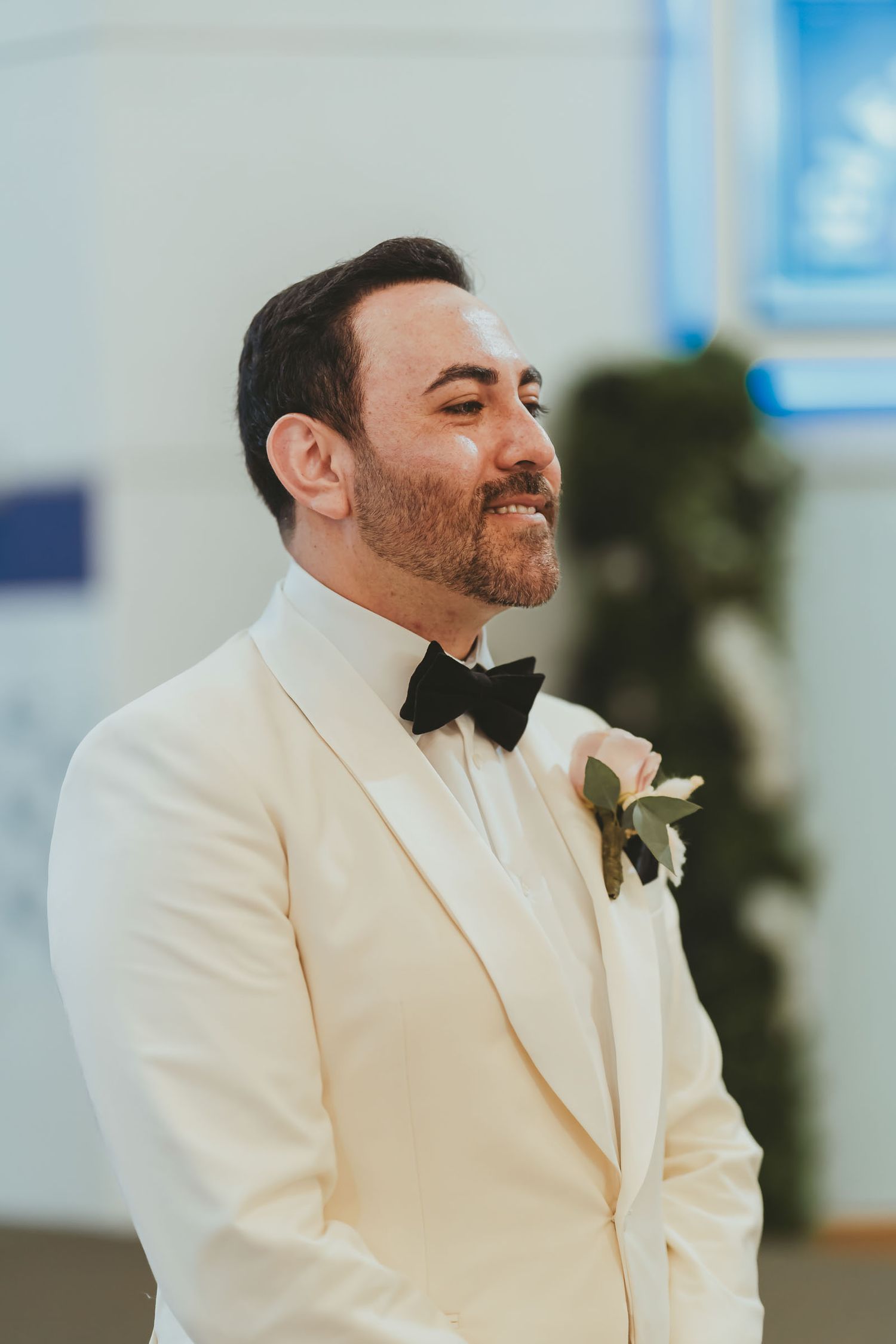 Novio con traje blanco y pajarita negra espera sonriente en altar durante ceremonia boda.