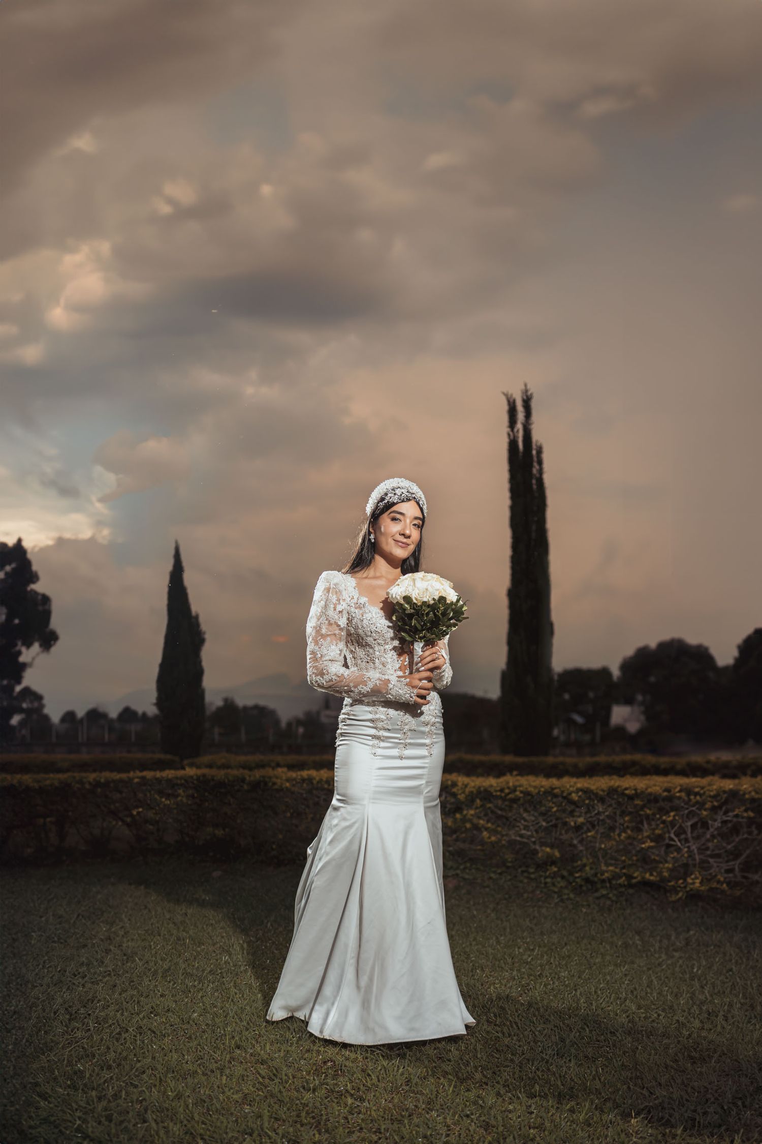 Novia con vestido sirena y ramo posa en exterior bajo cielo nublado dramático; árboles de pinos al fondo.