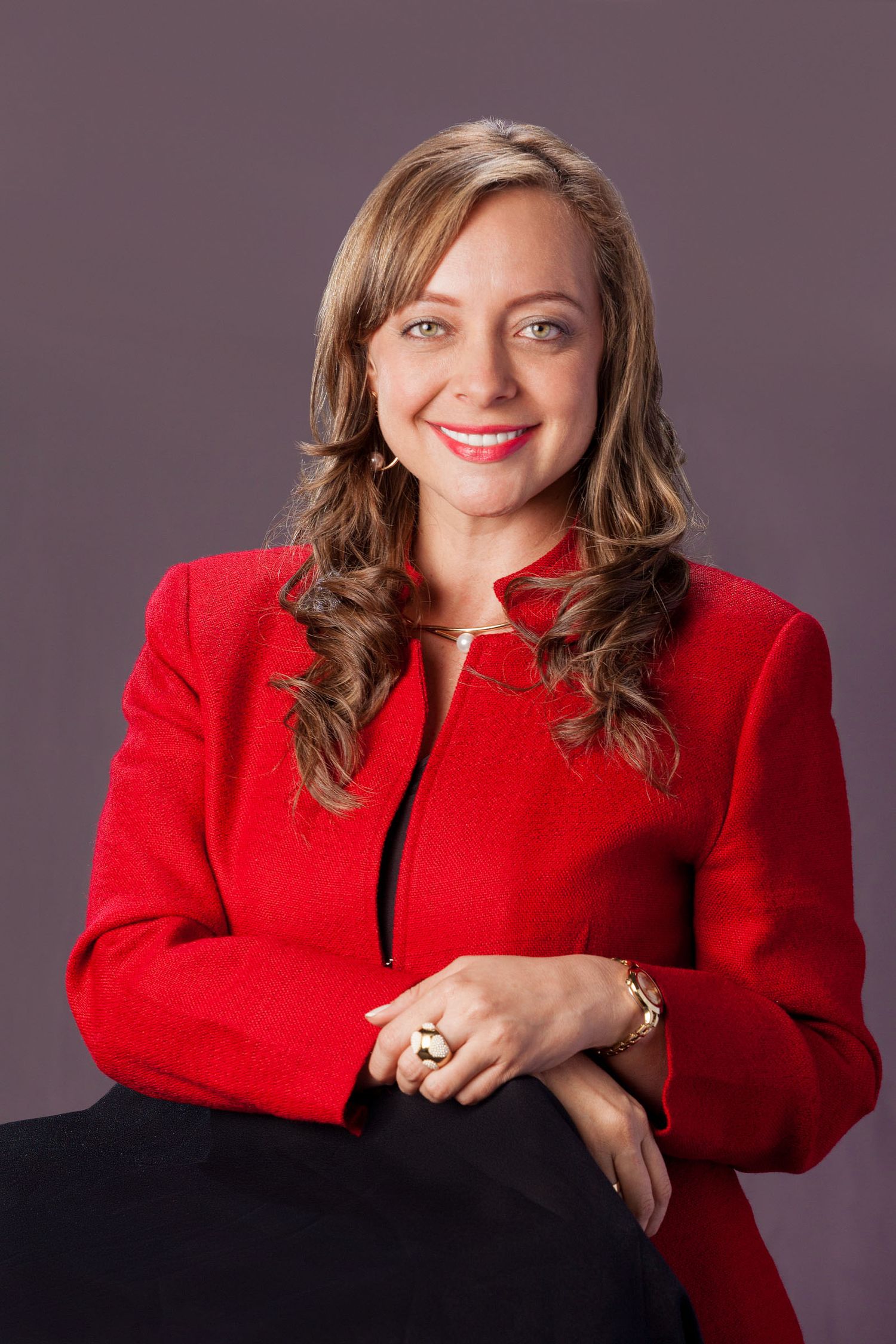 Retrato mujer sonriente con blazer rojo vibrante sobre ropa oscura, inclinada hacia adelante; fondo gris estudio. Ana Paola Agudelo