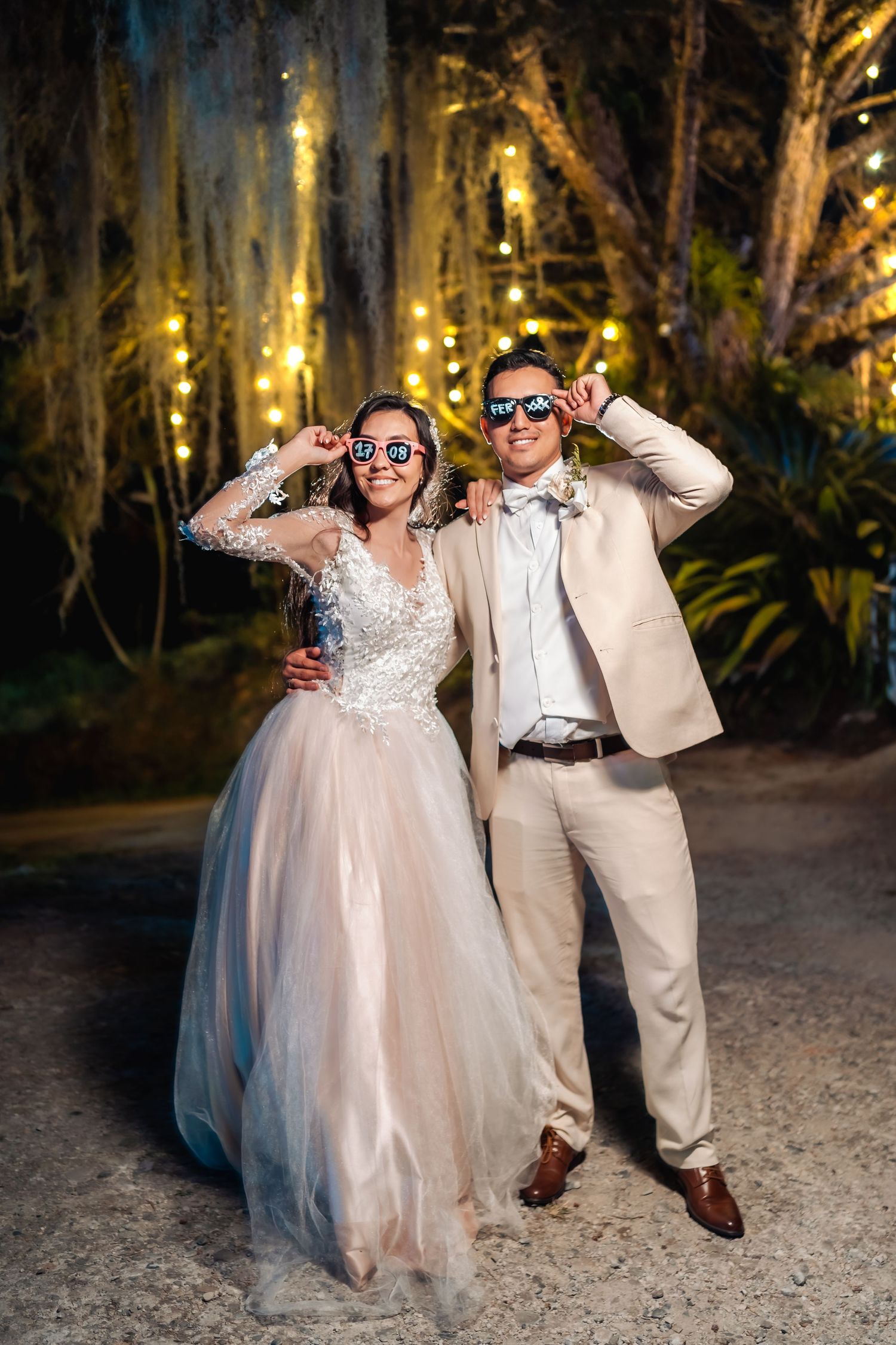 Novios posan con gafas de fiesta en recepción nocturna; ella vestido tul, él traje claro. Boda divertida.