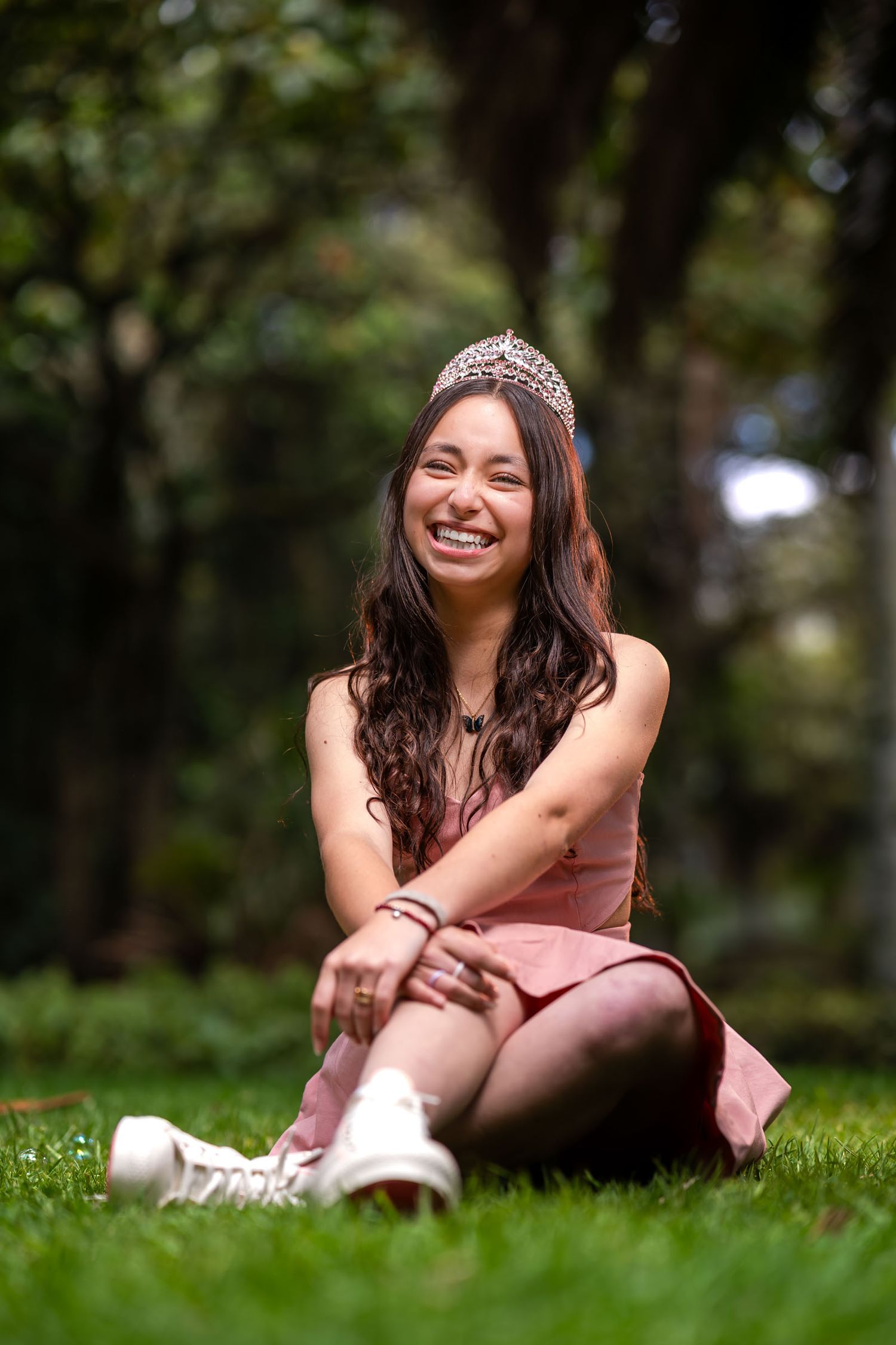Joven quinceañera sonriente con vestido corto rosa, tiara y zapatillas, sentada en césped; sesión prequince.