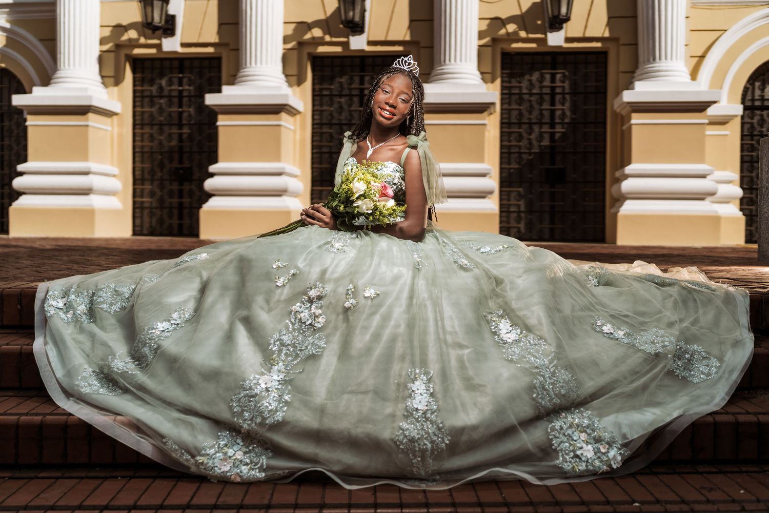 Quinceañera afrodescendiente sonríe sentada en escaleras con vestido princesa verde/plata extendido; edificio clásico.