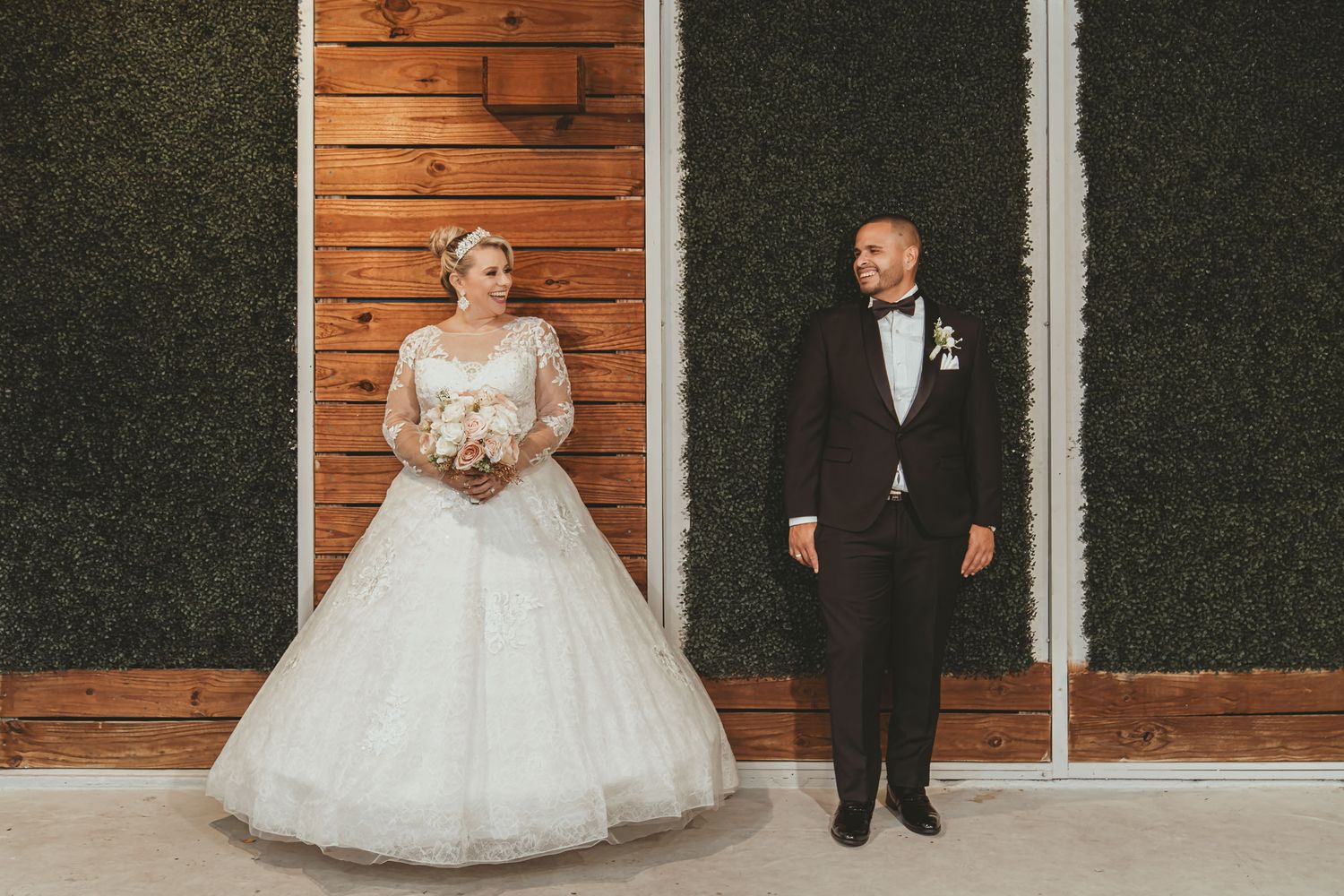 Novios se miran posando separados frente a pared moderna (madera y follaje); ella sostiene ramo. Foto boda.