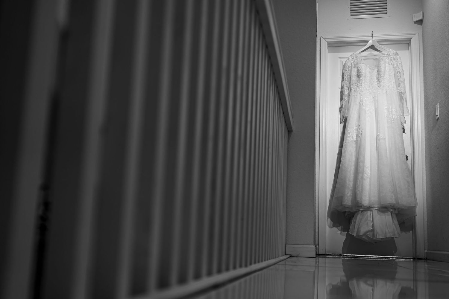 Foto B&W: Vestido de novia colgado en puerta al final de pasillo; barandal desenfocado primer plano. Boda.
