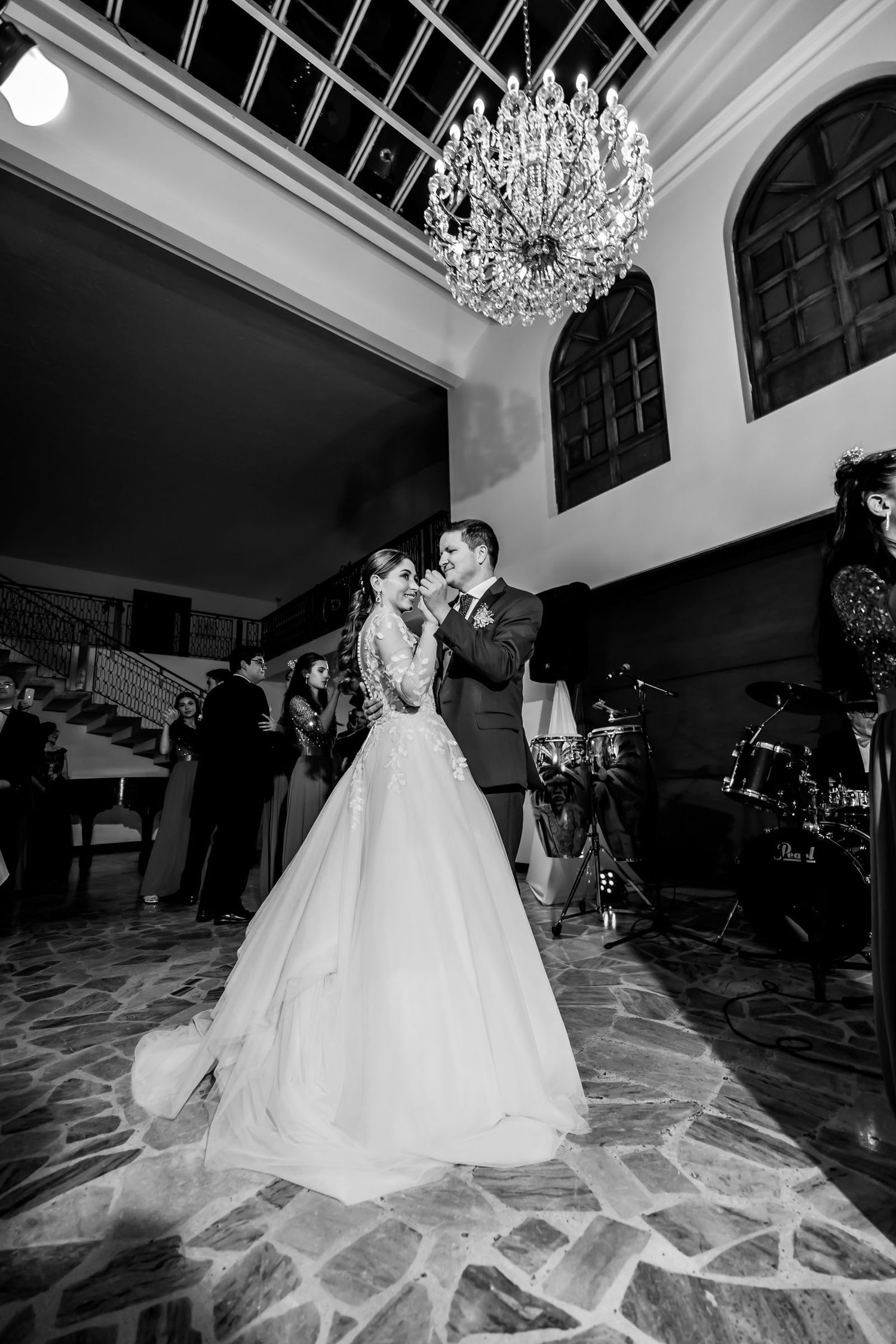 Foto B&W: Novios comparten primer baile bajo gran candelabro en salón de recepción; fotografía de boda elegante.