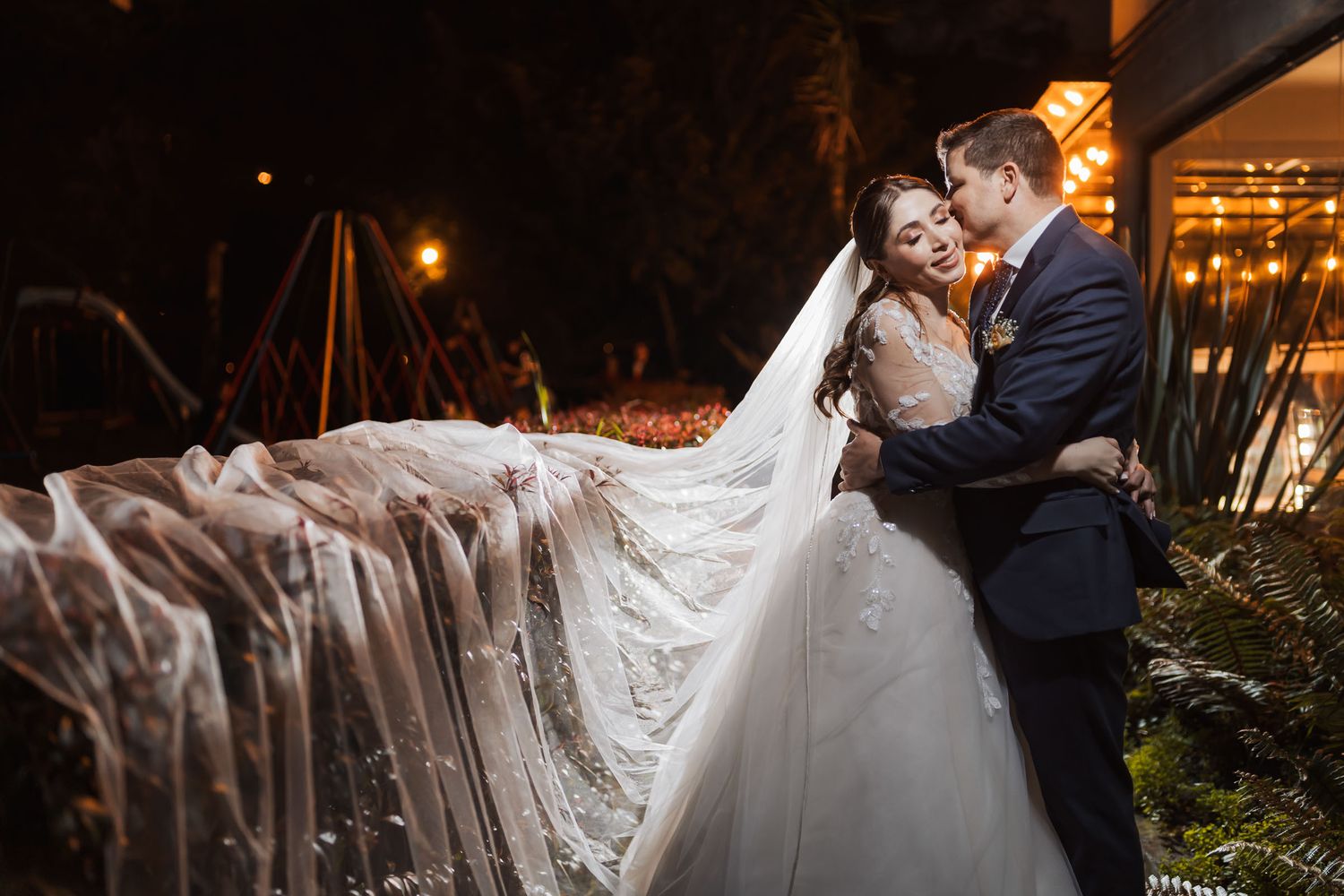 Novio abraza a novia sonriente por detrás; su velo largo se extiende dramáticamente. Foto nocturna boda exterior.